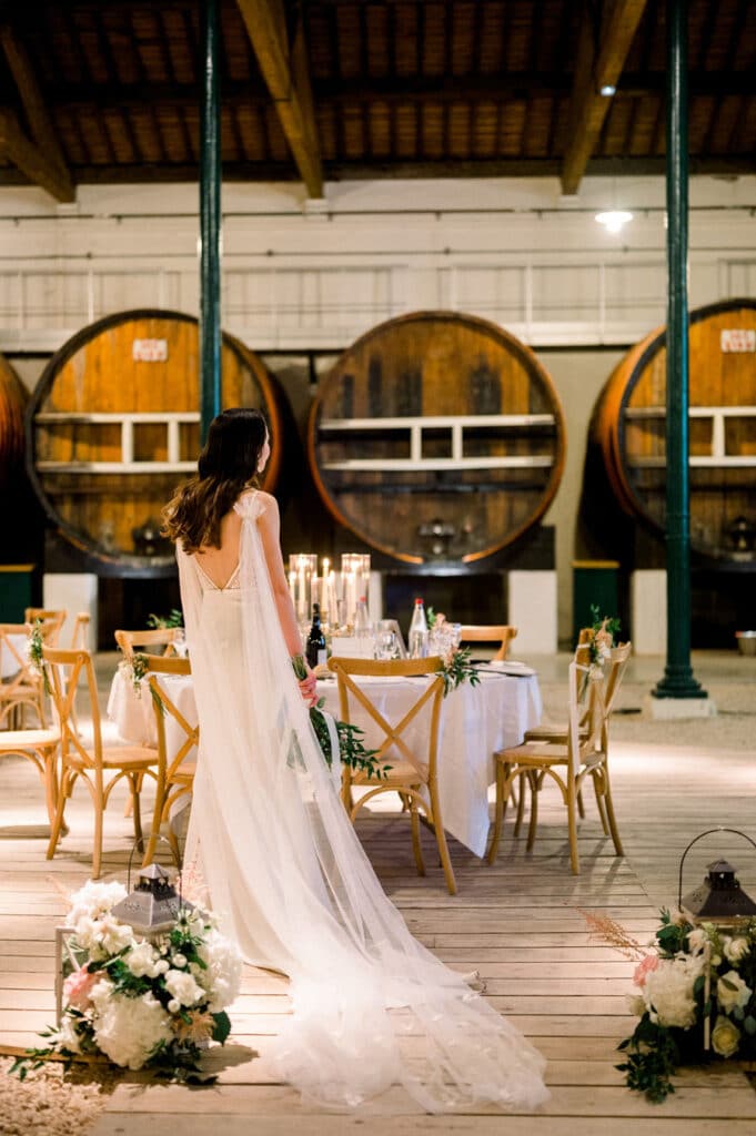 Mariée en cape blanche devant de grands tonneaux de vin. Réception de mariage élégante dans un chai, décorée de bougies et fleurs.