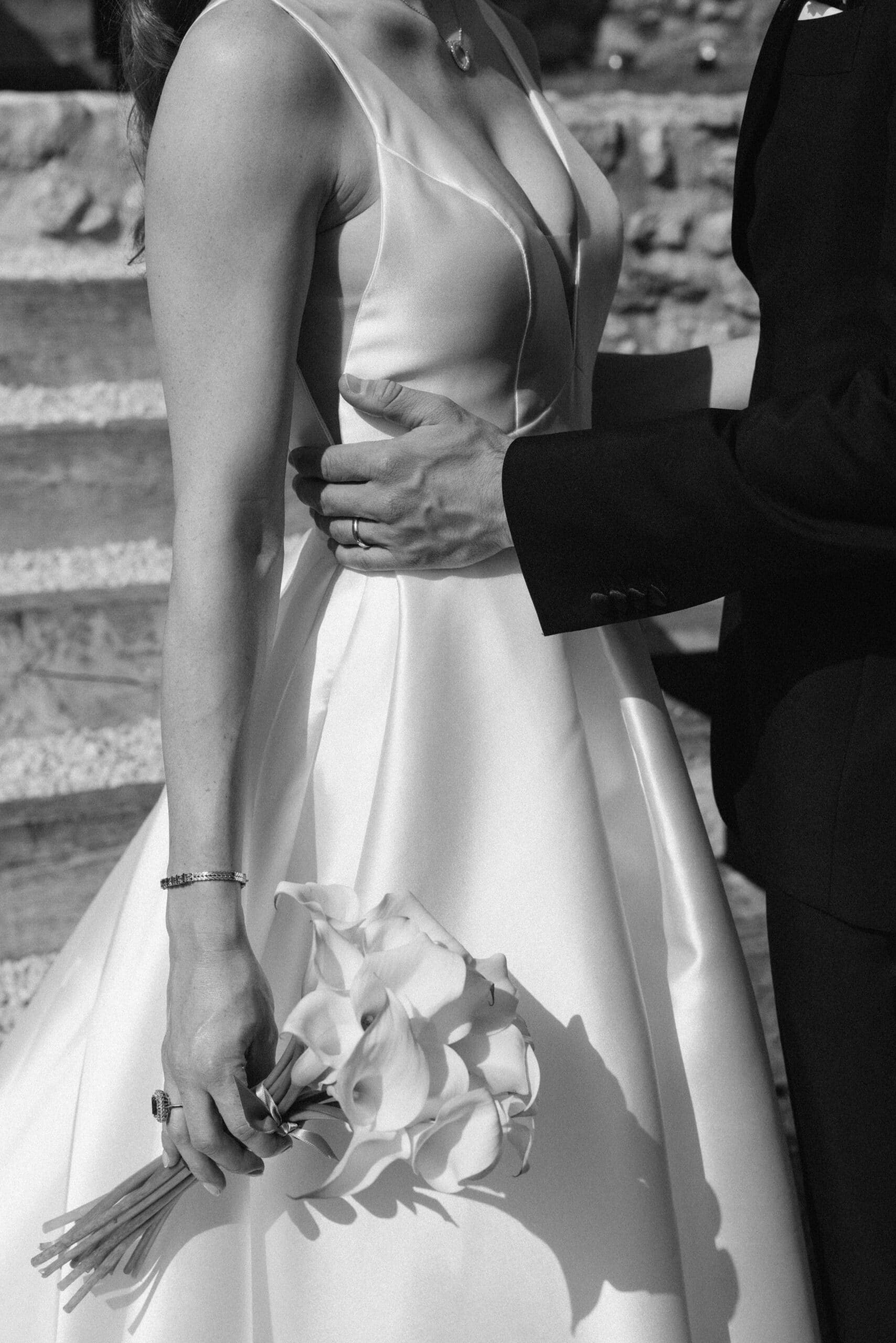 Noir et blanc : Main du marié avec alliance ceinturant la taille de la mariée en robe de satin. Elle tient un bouquet de callas.