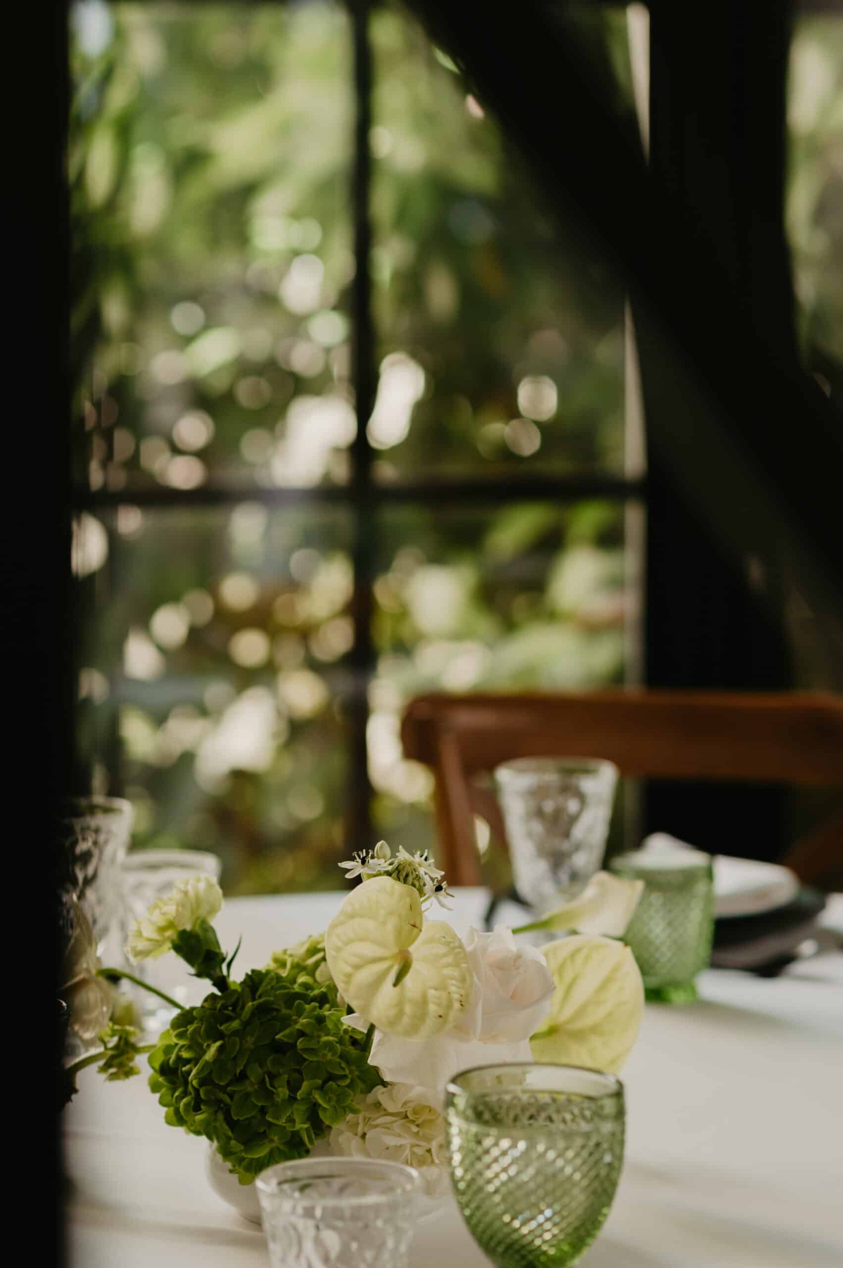 Centre de table frais: fleurs vertes et blanches (anthurium), verres texturés verts. Ambiance événementielle avec un fond de jardin lumineux (bokeh).
