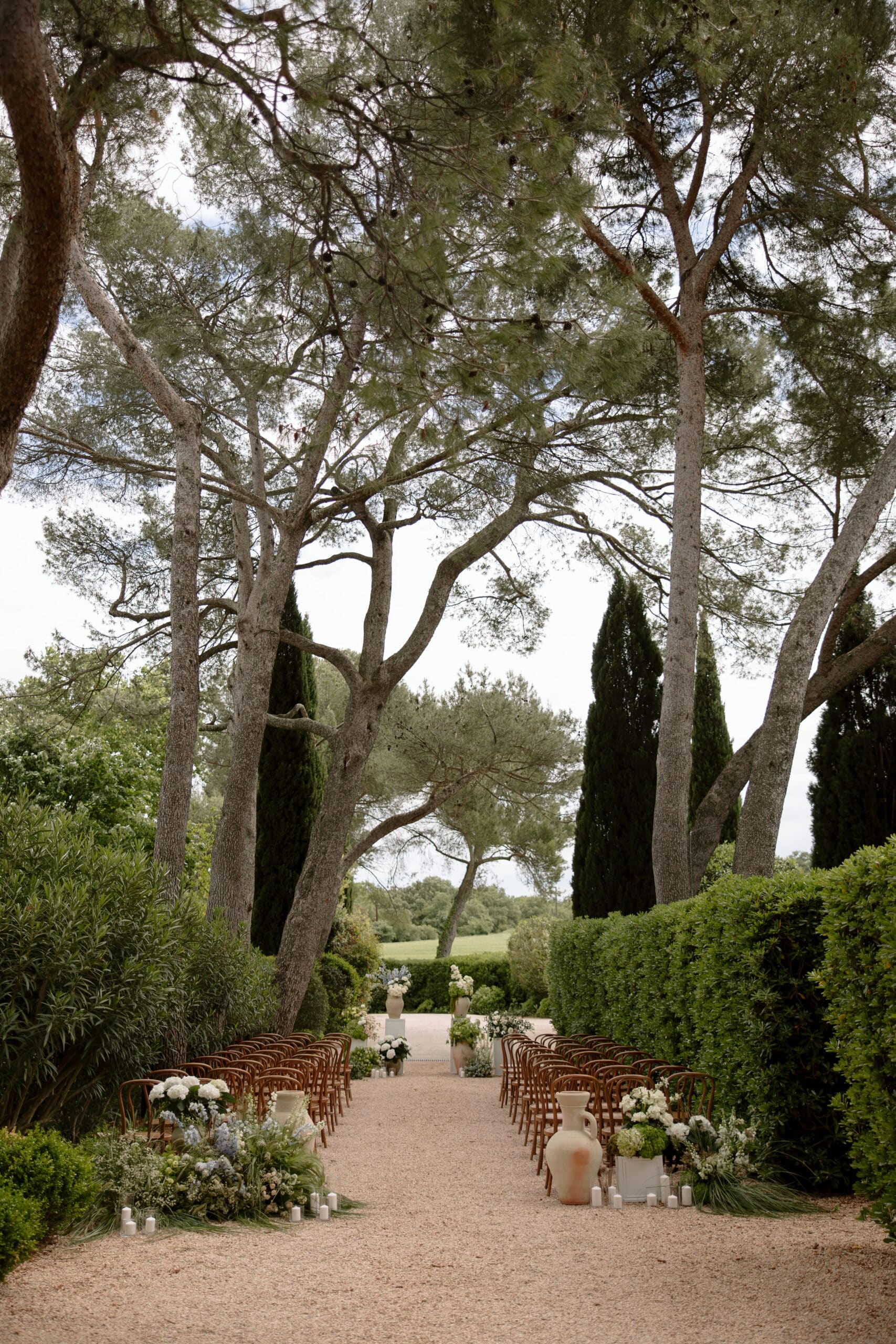 Allée de cérémonie de mariage en gravier bordée de chaises en bois et de compositions florales bleues et blanches sous les grands arbres.