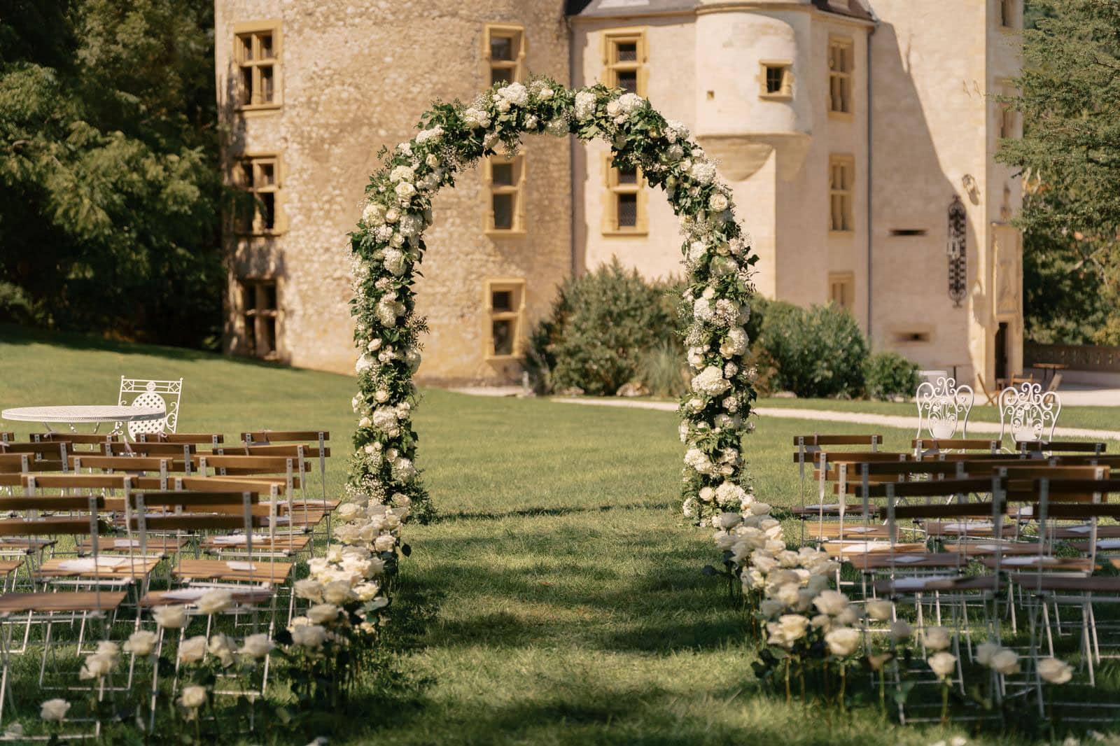 Cérémonie de mariage extérieure devant un château. Arche florale massive en roses blanches et allée bordée de fleurs pour les invités.