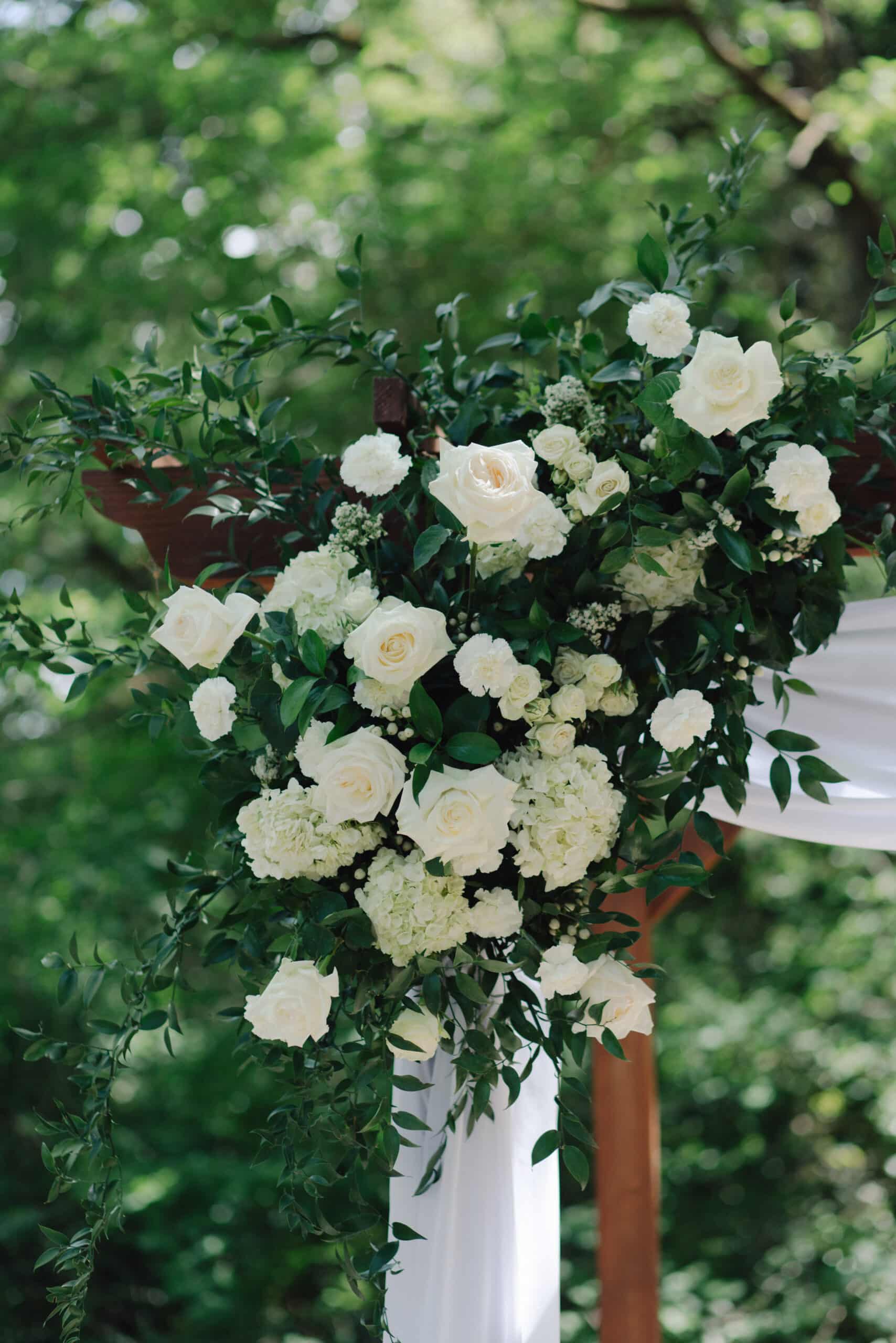 Luxueux arrangement floral d'arche de mariage: roses et hortensias blancs, verdure luxuriante (ruscus) et drapé de tissu blanc.