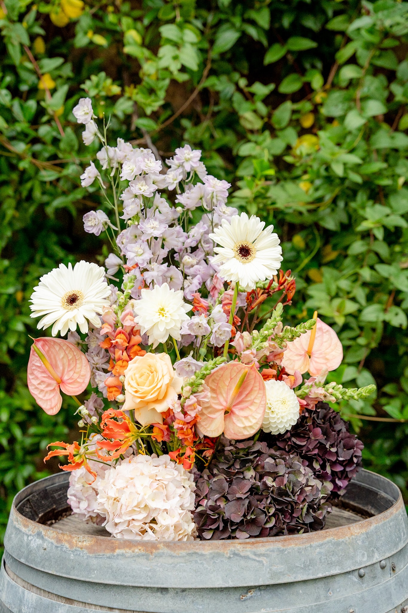 Bouquet rustique en tons pêche et rouille: Gerbera, Anthurium, roses, delphinium lavande et hortensia sombre, dans un fût en métal.