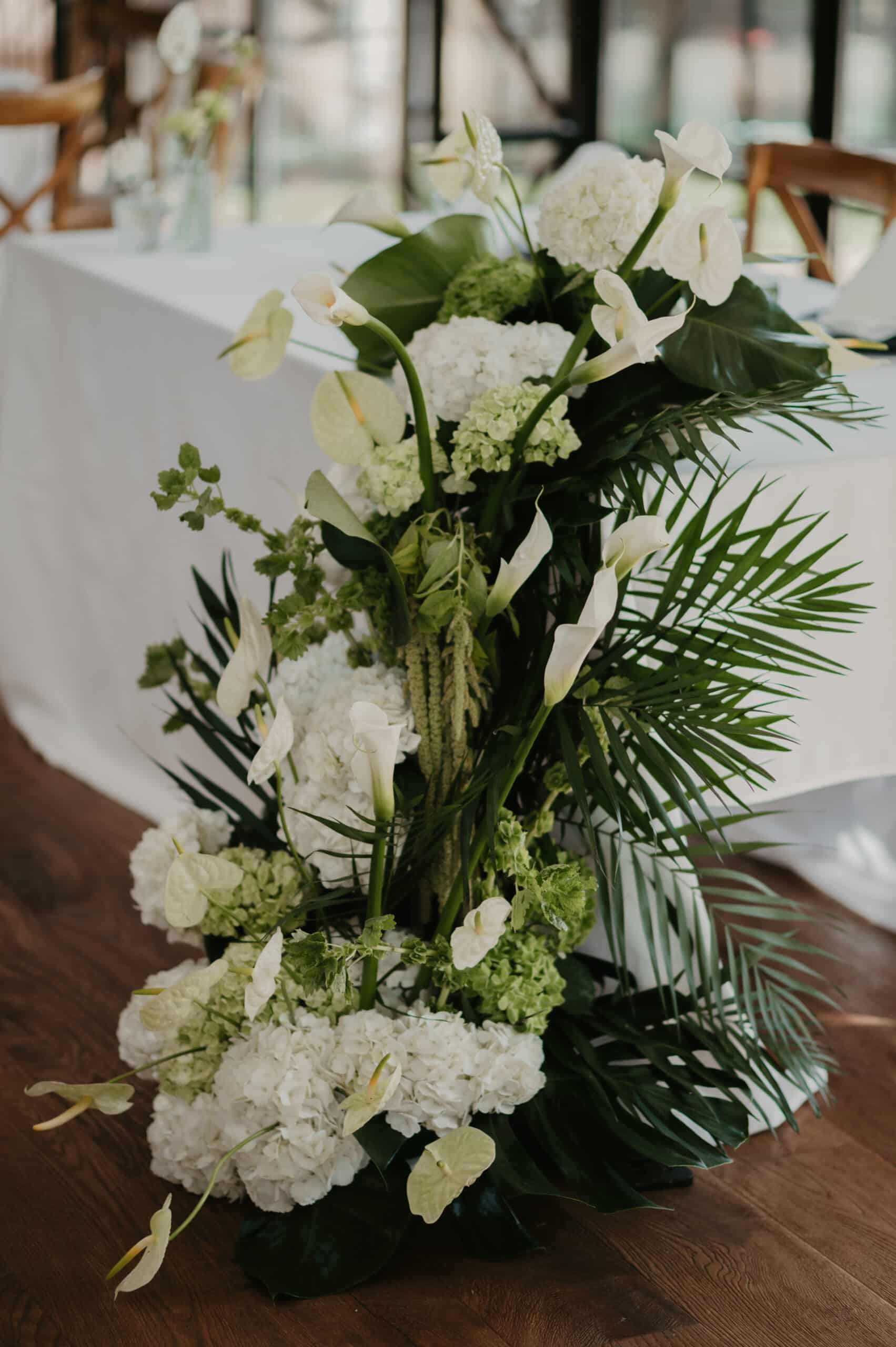 Lush arrangement floral blanc/vert pour événement : hydrangées, callas, anthuriums et feuillage tropical près d'une table.