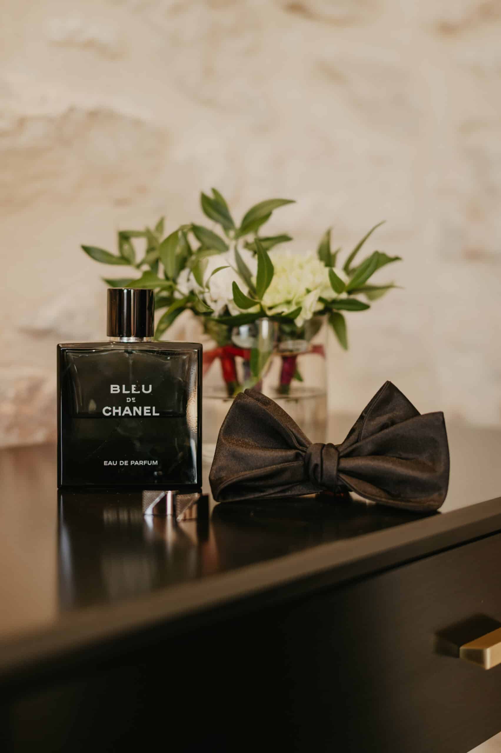 Bouteille Bleu de Chanel Eau de Parfum et nœud papillon noir sur meuble sombre. Bouquet flou en fond. Ambiance élégante.