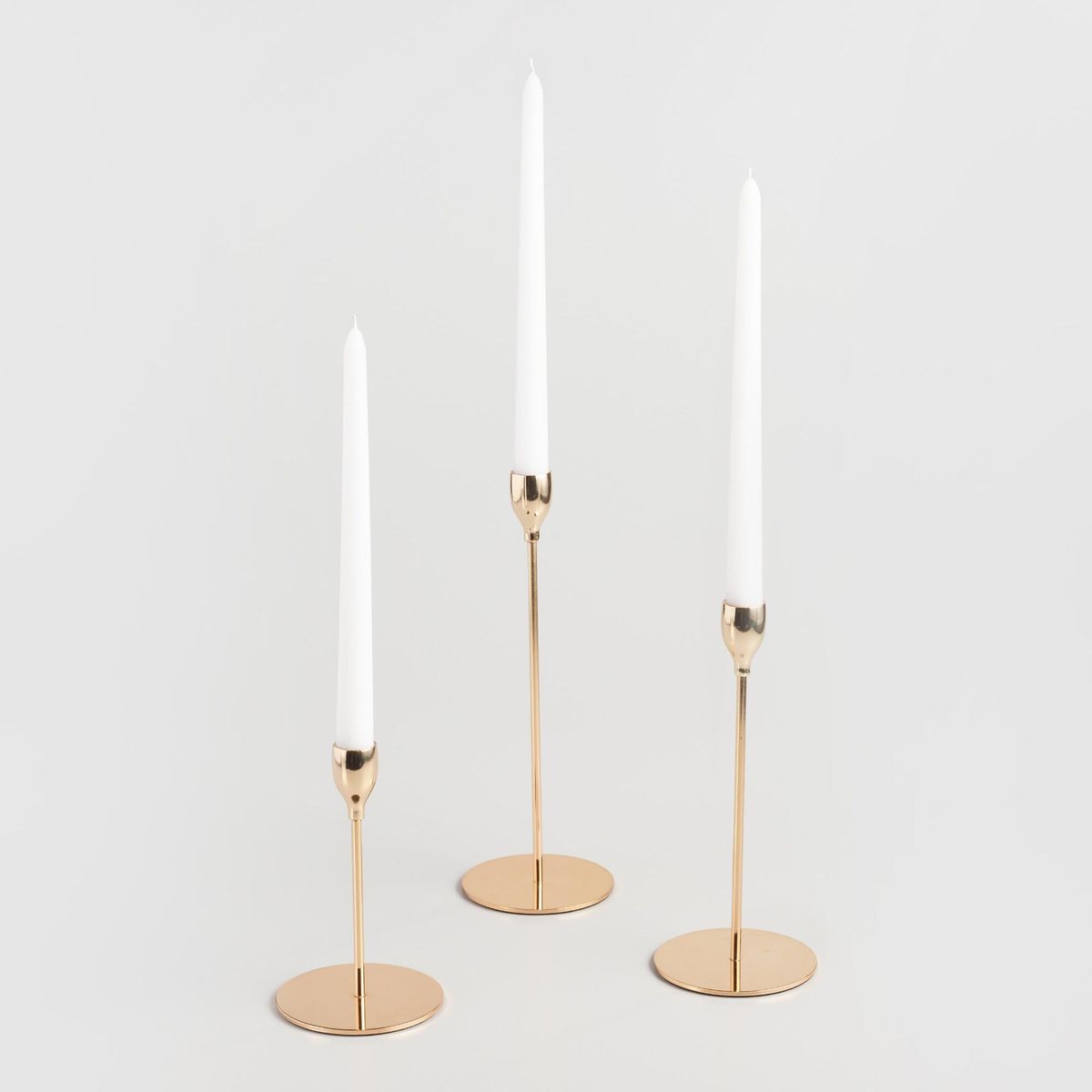 Trois élégants bougeoirs minimalistes en or avec de longues chandelles coniques blanches, posés sur un fond clair.