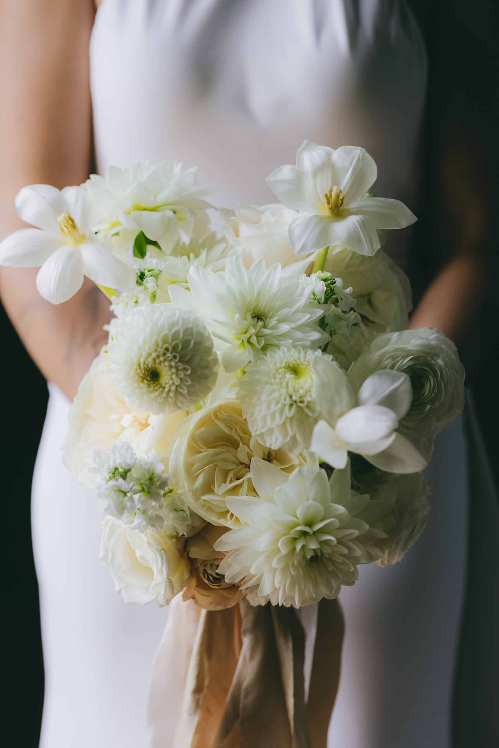 Sublime Bouquet de Mariée Blanc : Dahlias et Roses Crème. Élégant bouquet de mariée crème et blanc contre une robe minimaliste. Mélange de dahlias, roses et ranunculus, lié par un ruban de soie.