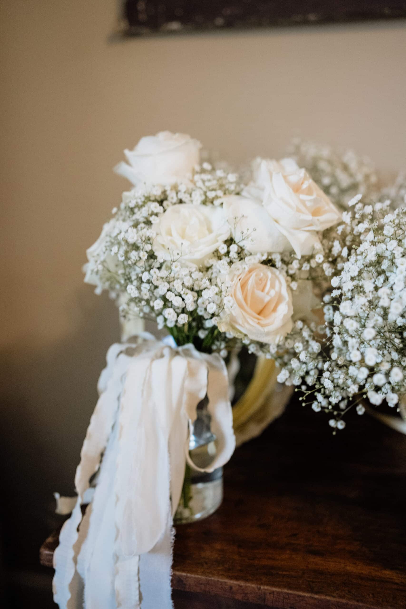 Bouquet de mariage romantique: roses ivoire et gypsophile dans un vase, ornés de longs rubans rustiques sur bois foncé.