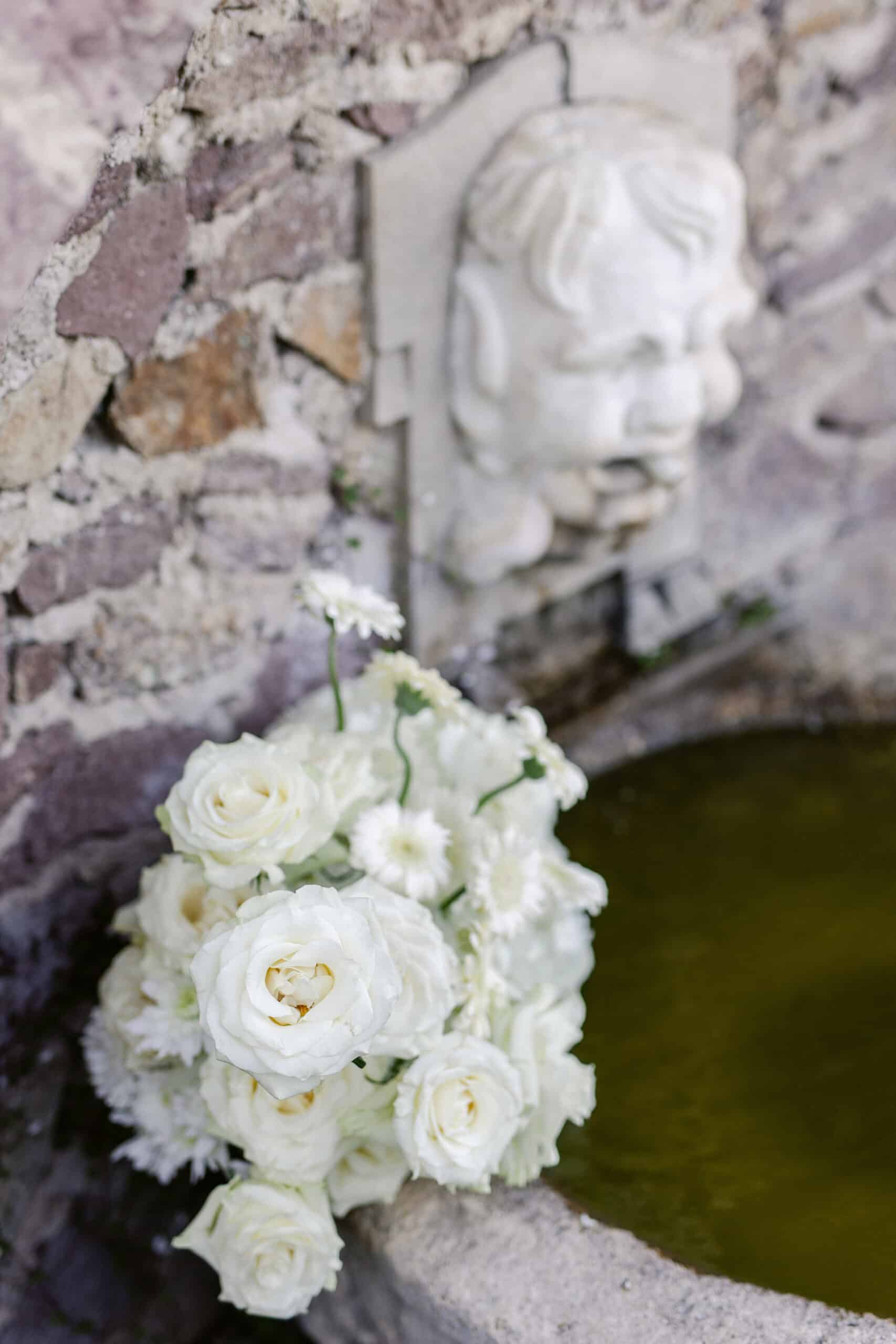 Magnifique bouquet de roses blanches sur le bord d'une fontaine en pierre, près d'un mur rustique orné d'une tête sculptée en marbre.