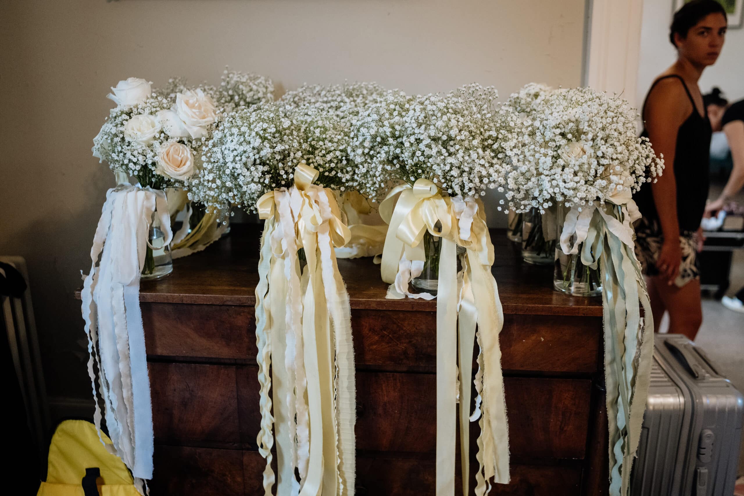 Bouquets de mariée gypsophile et roses sur commode en bois, décorés de longs rubans crème. Une femme observe lors des préparatifs.