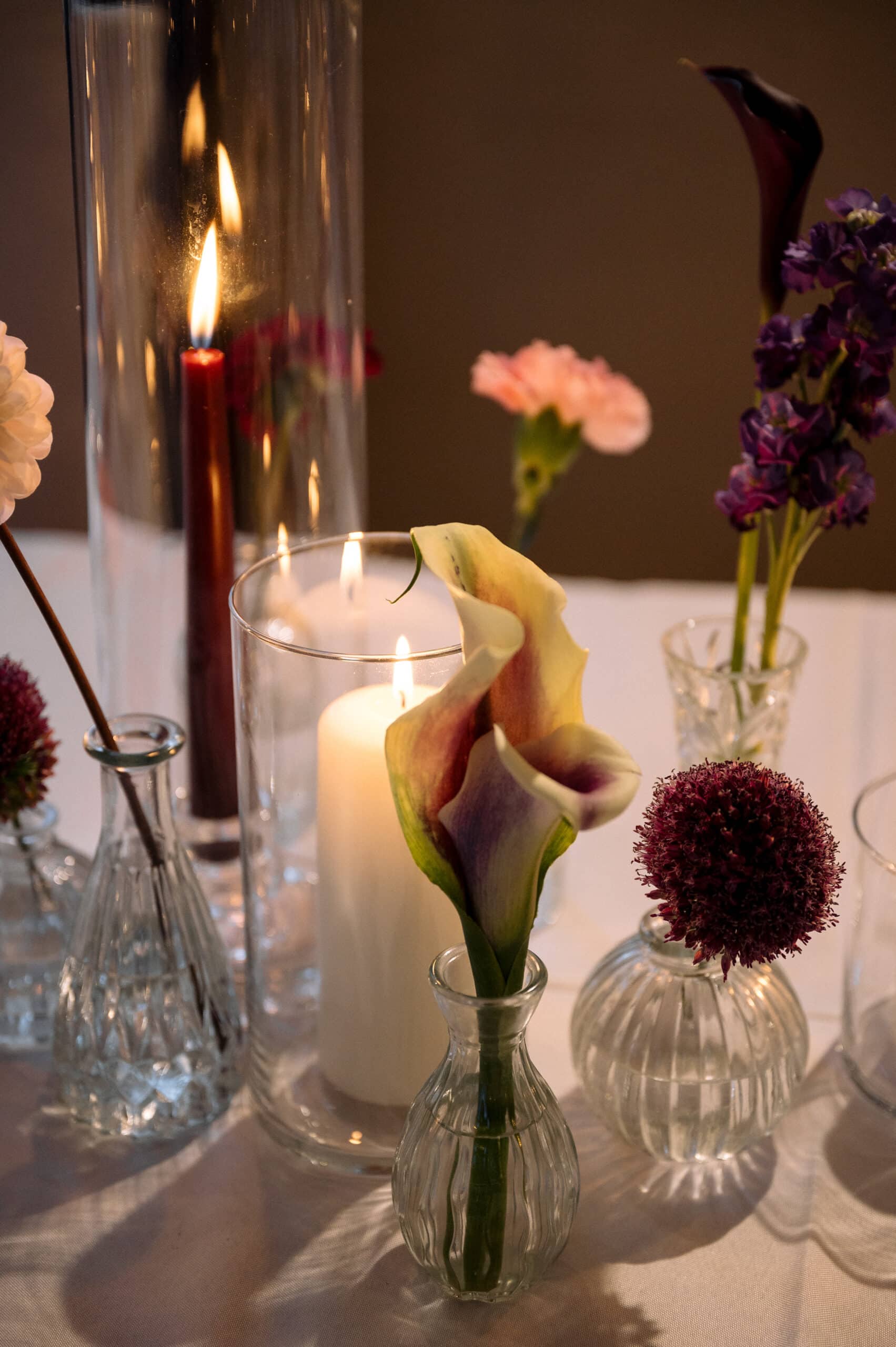 Centre de table intime: bougies rouges et blanches dans des verres, entourées de petits vases de fleurs, dont un arum jaune et un allium foncé.