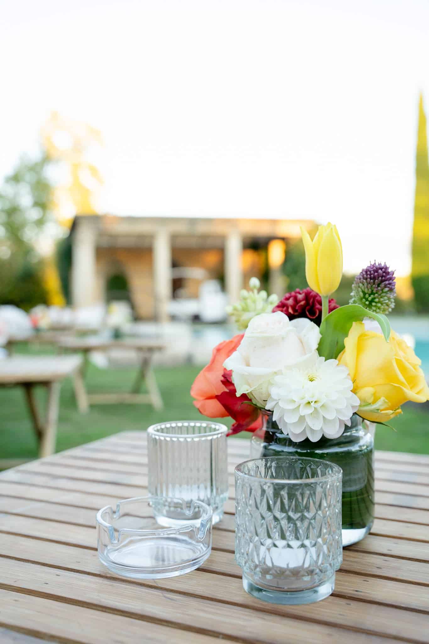 Bouquet de fleurs colorées (tulipe jaune, rose, dahlia) sur table en bois avec verres et cendrier. Décoration de réception en extérieur.