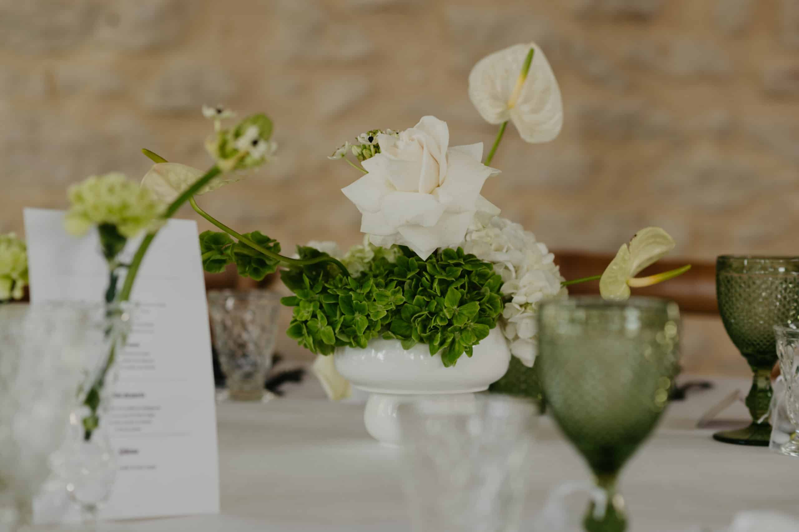 Élégant centre de table de mariage : rose blanche, anthuriums crème et verdure d'hortensia. Vase blanc sur nappe; vaisselle verte.