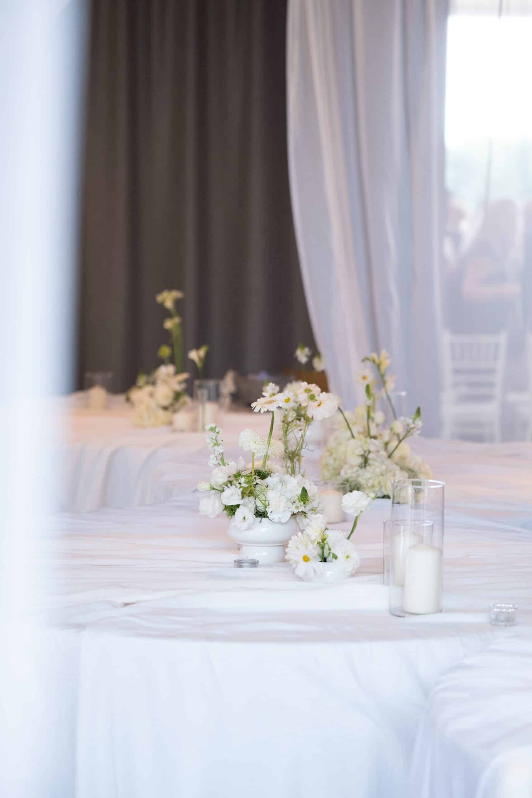 Élégant centre de table de réception avec compositions florales blanches (dahlias, marguerites) et bougies sur nappes immaculées.