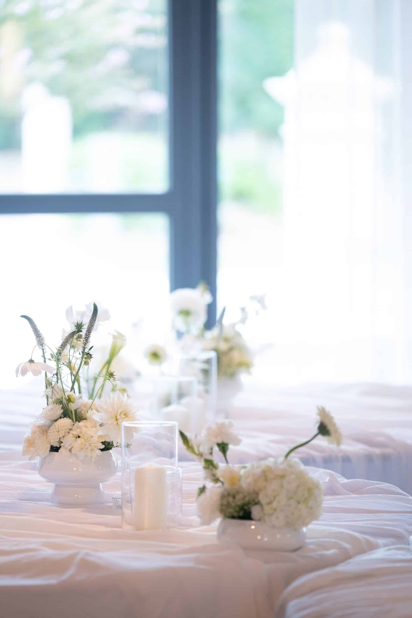 Centres de table mariage élégants: arrangement de fleurs blanches, dahlias et bougies sur nappe drapée, près d'une fenêtre lumineuse.
