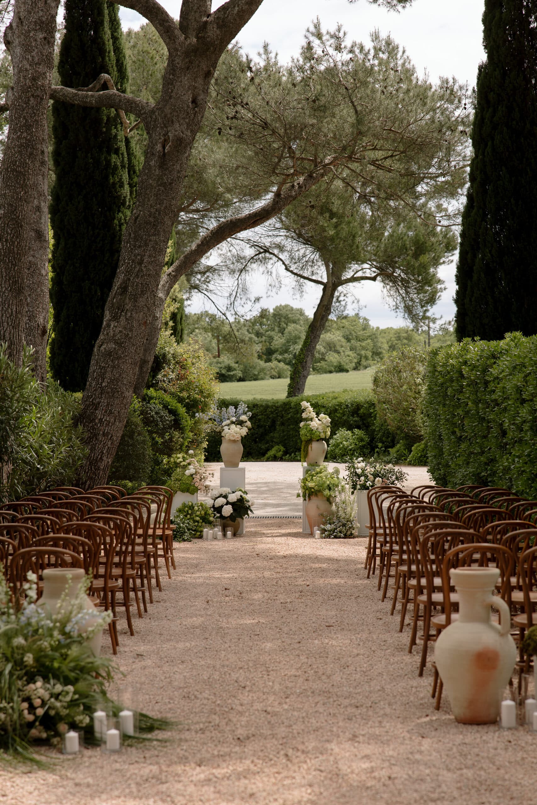 Longue allée de mariage en gravier bordée de chaises Thonet. Urnes antiques et compositions florales blanches pour la cérémonie en jardin.