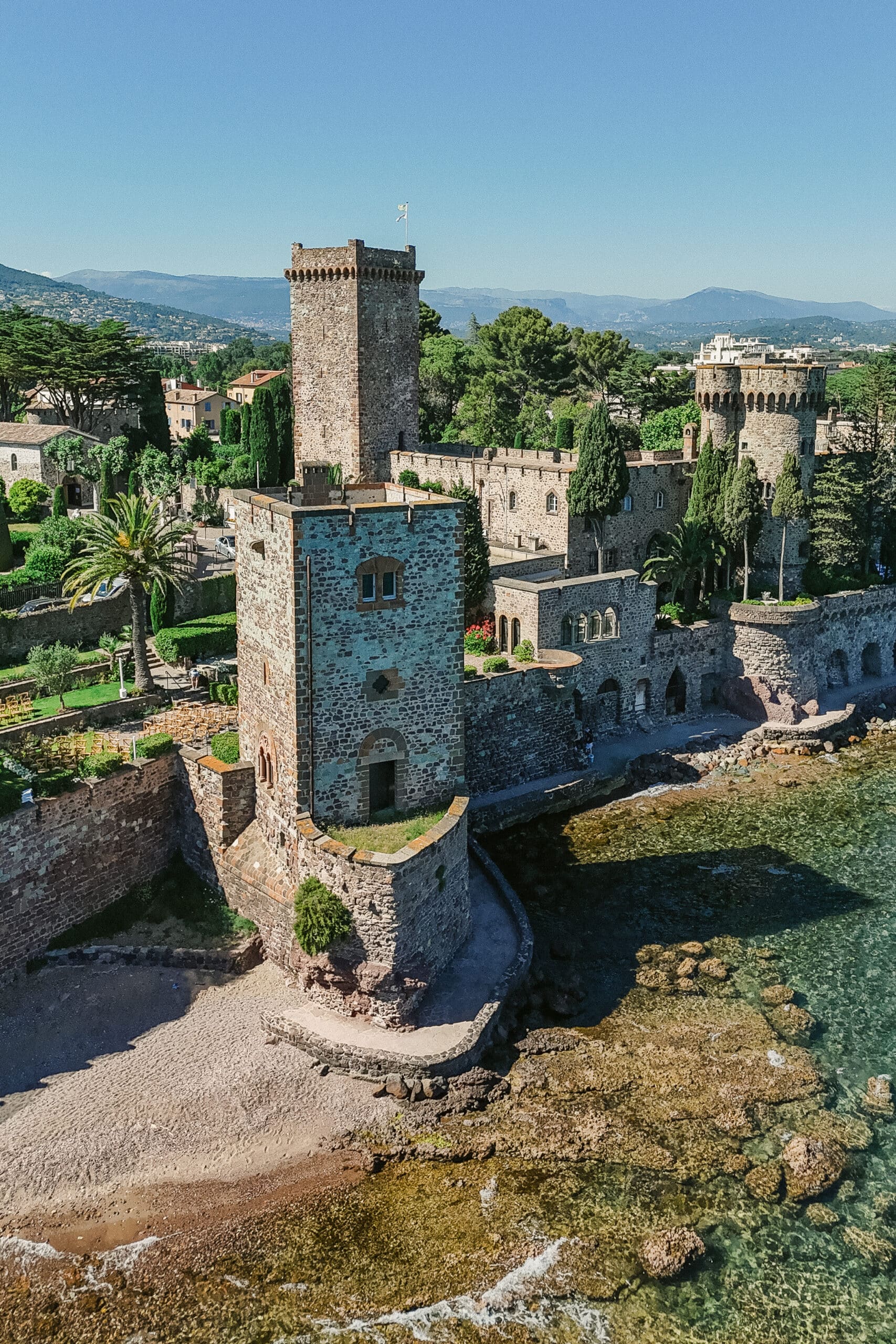 Imposant château médiéval en pierre rouge sur la côte Méditerranéenne. Jardins luxuriants, tours crénelées et plage rocheuse.