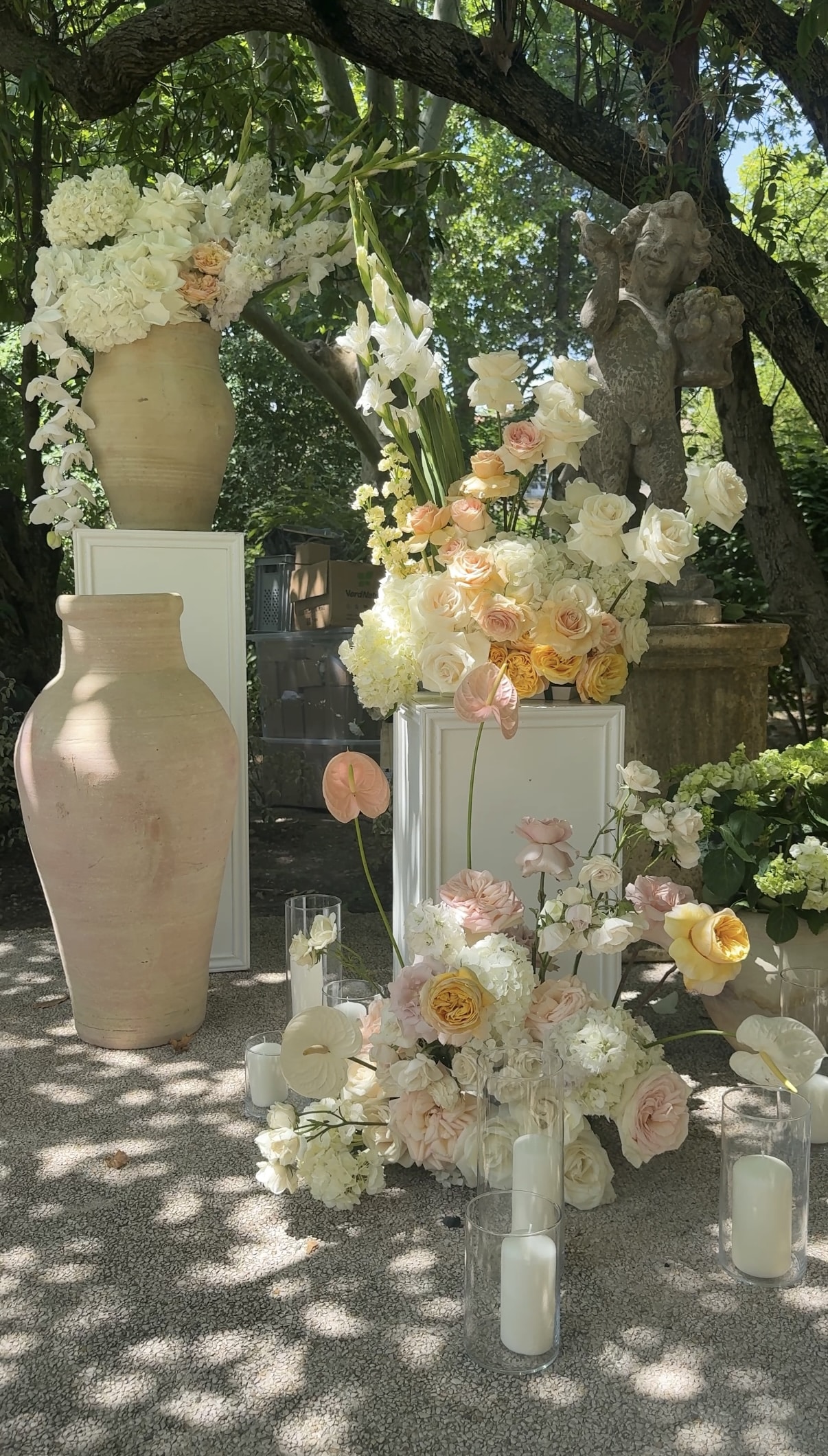 Luxe composition florale de mariage en extérieur : roses pastel, hydrangées, anthuriums, vases terre cuite, socles blancs et bougies.