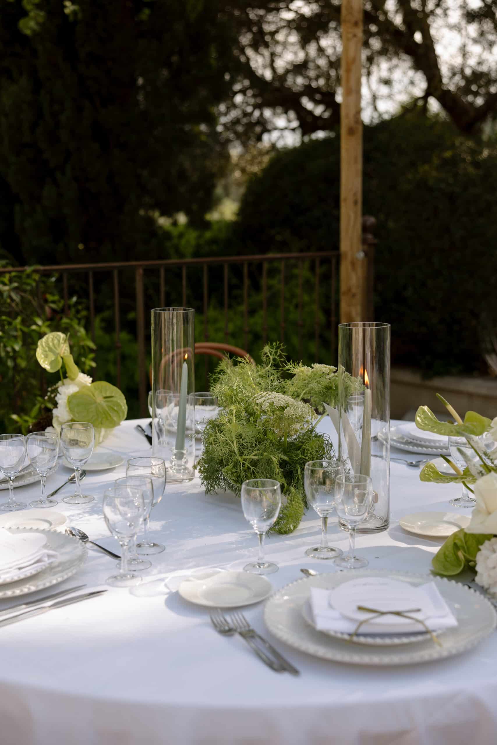 Table de mariage élégante dressée en extérieur. Centre de table végétal vert et blanc, bougies sauge sous verre, vaisselle nacrée.
