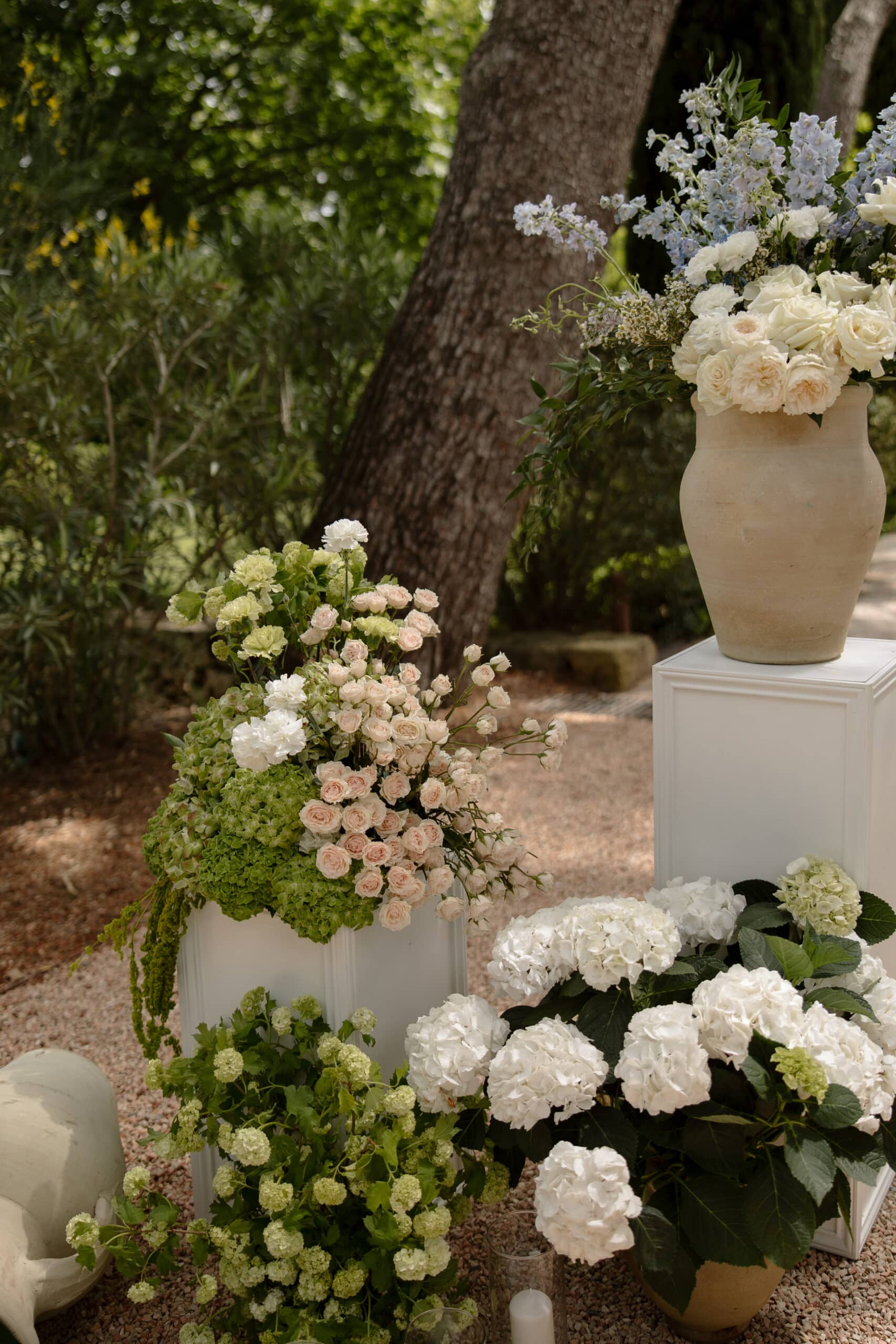 Bouquet de roses crème, d'hortensias verts et de delphiniums bleus, présentés dans des urnes et des piédestaux blancs, décorant un jardin ombragé.
