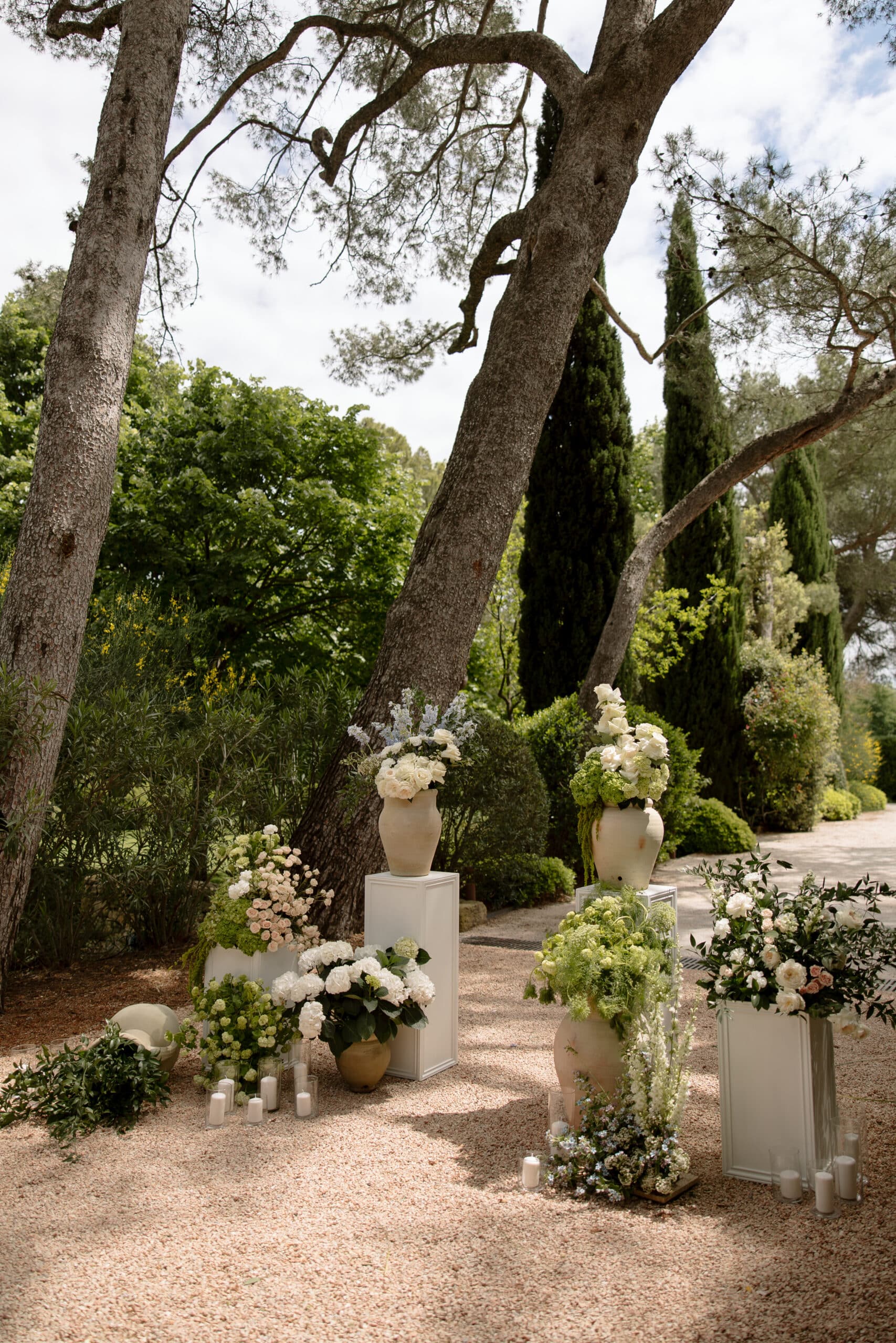 Installation florale de mariage en extérieur : roses blanches, hortensias et delphiniums bleus dans de grands vases sur socles blancs, sous un pin.