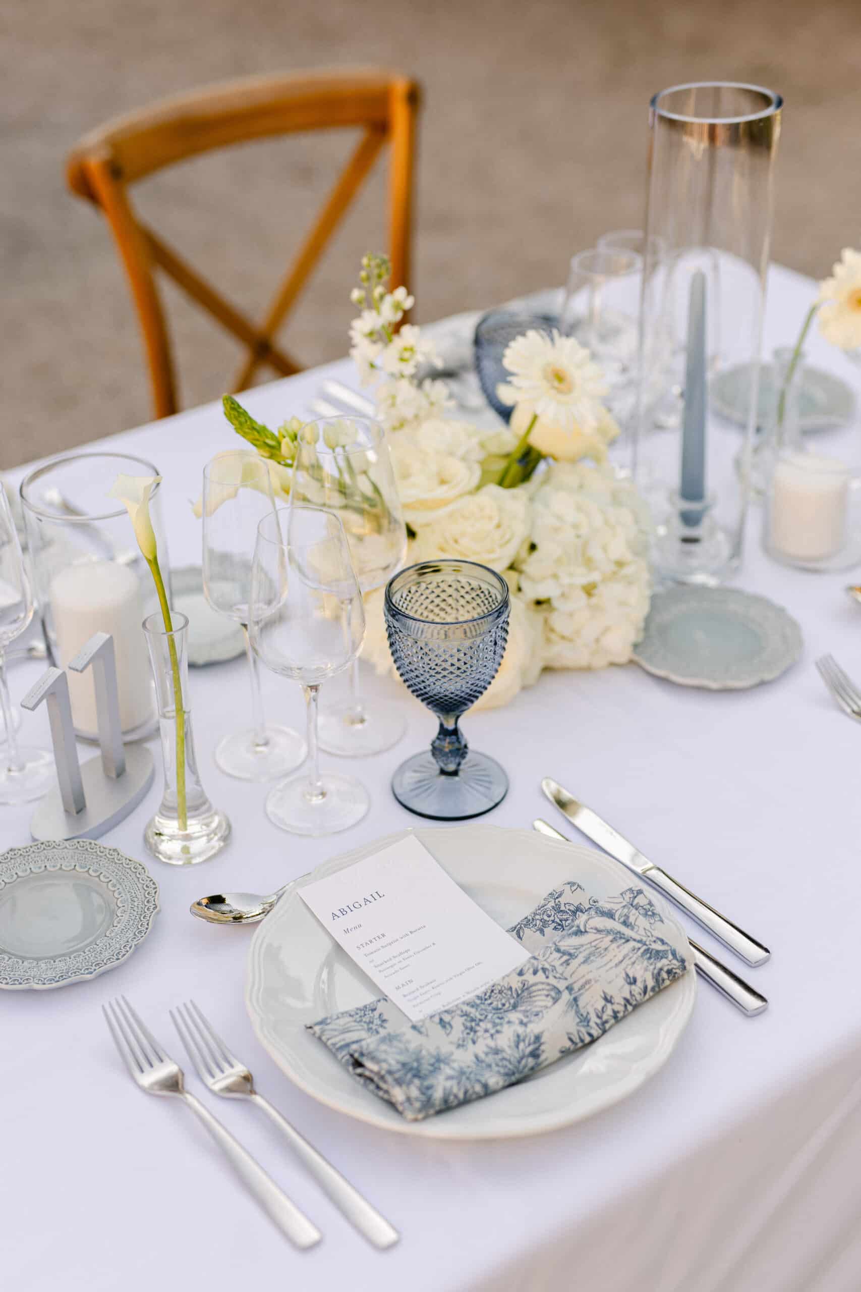 Table de mariage extérieure: centre de roses blanches, goblet bleu, bougies grises. Menu u0022ABIGAILu0022, serviette toile de Jouy et couvert argenté.