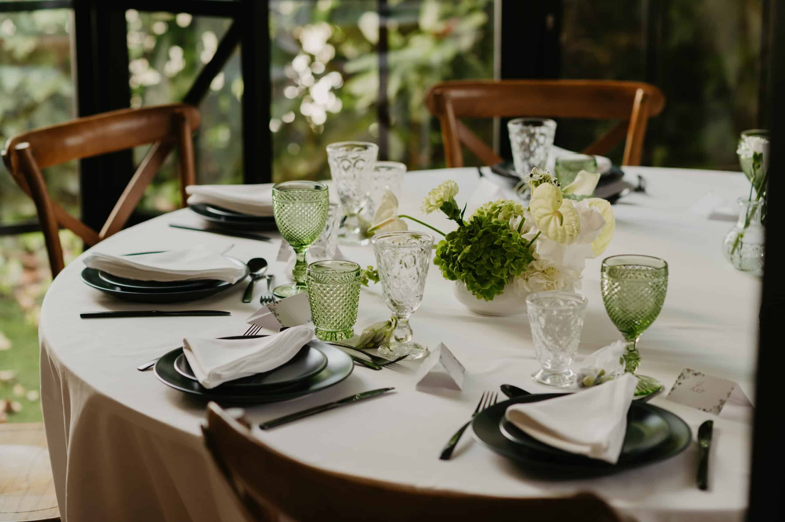 Table de mariage élégante: nappage blanc, vaisselle vert foncé et gobelets vintage. Centre floral luxuriant devant un jardin lumineux.