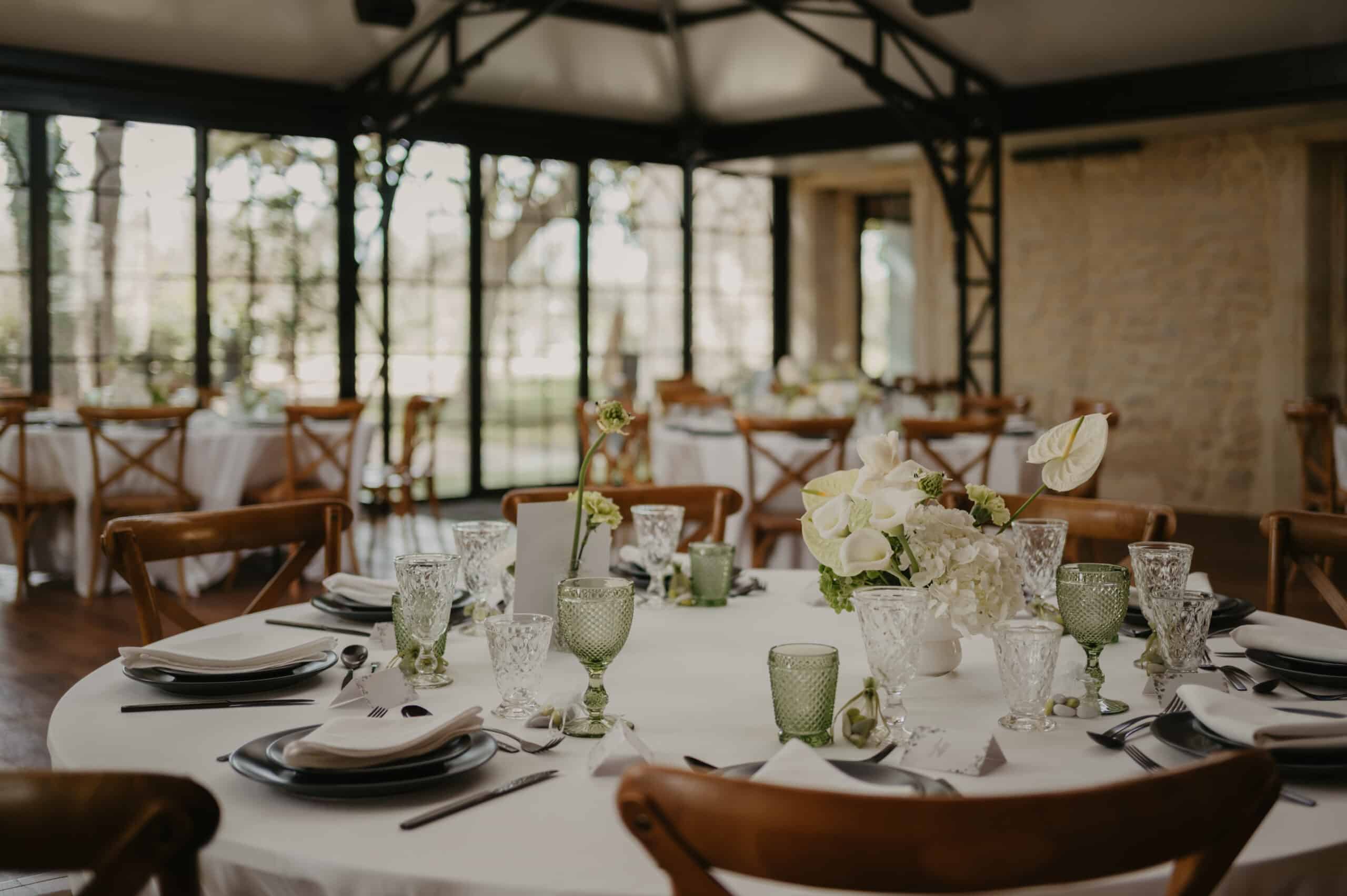Table de mariage dans un pavillon vitré. Décoration chic: verres verts, vaisselle noire, chaises X-back bois et centre de table floral blanc.