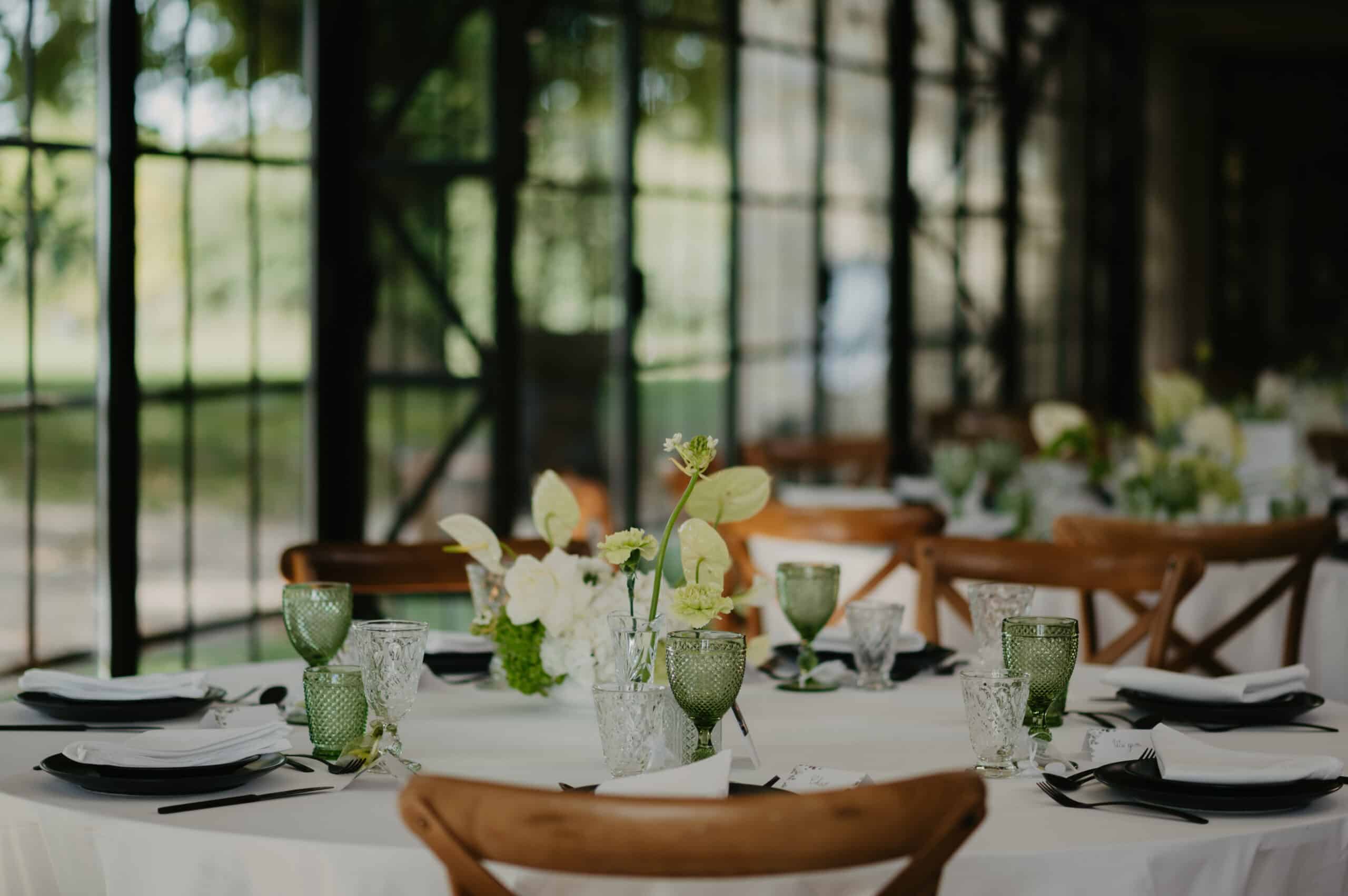Table de mariage élégante sous verrière : centre floral blanc et vert pâle, verres verts texturés et vaisselle noire sur nappe blanche, chaises bois rustiques.