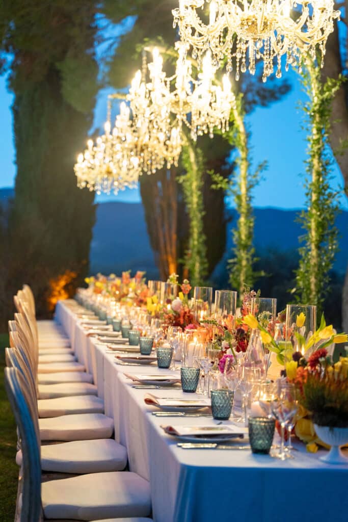 Longue table de réception extérieure illuminée par des lustres en cristal et des bougies. Décoration florale vive, verrerie bleue sur nappe blanche.
