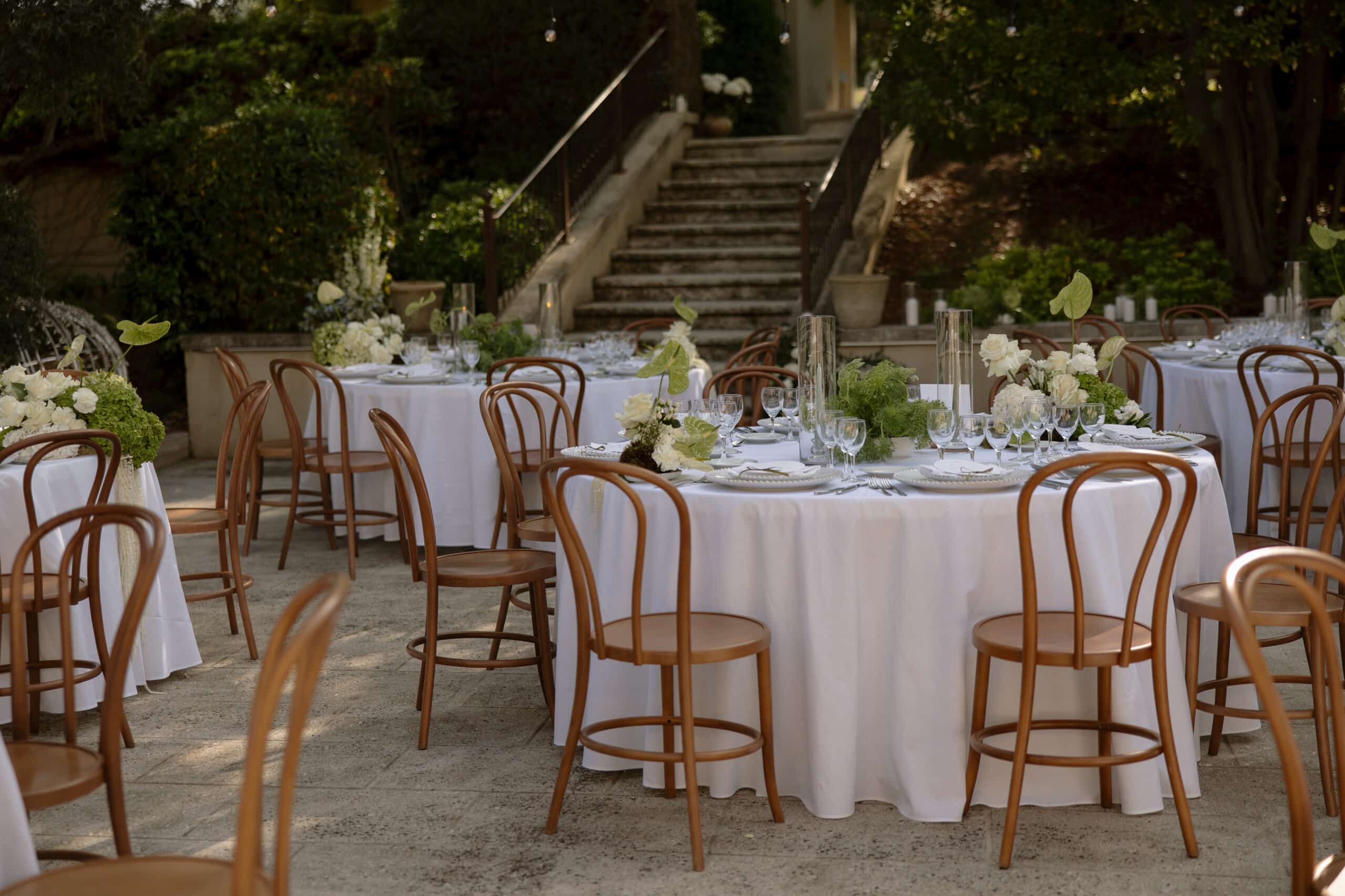 Réception extérieure élégante: tables rondes et chaises Thonet en bois sur une terrasse. Décor floral blanc/vert. Escalier en pierre à l'arrière.