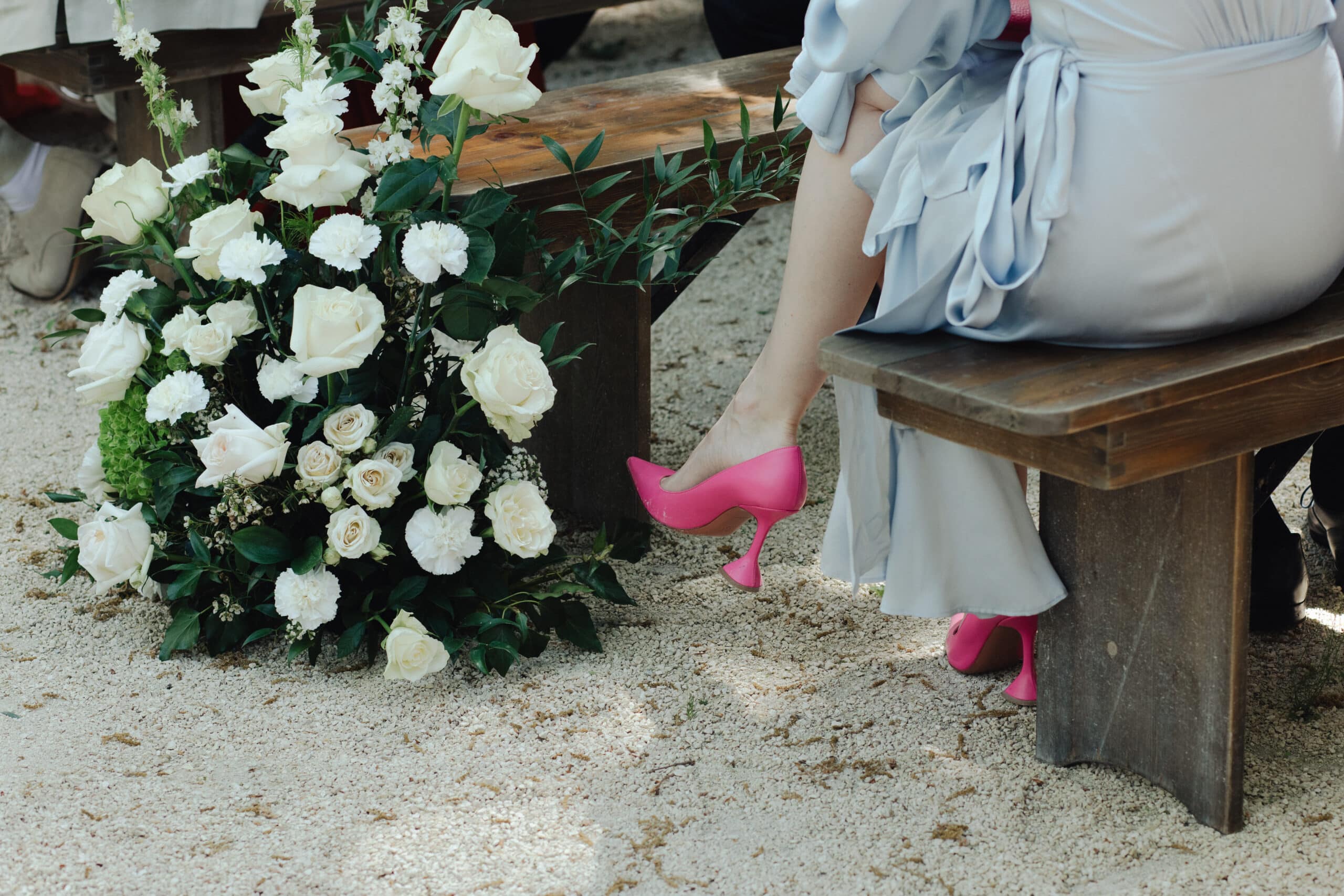 Escarpins fuchsia à talon sculpté sous une robe bleu ciel, assise sur un banc en bois. Grand arrangement de roses blanches sur gravier.