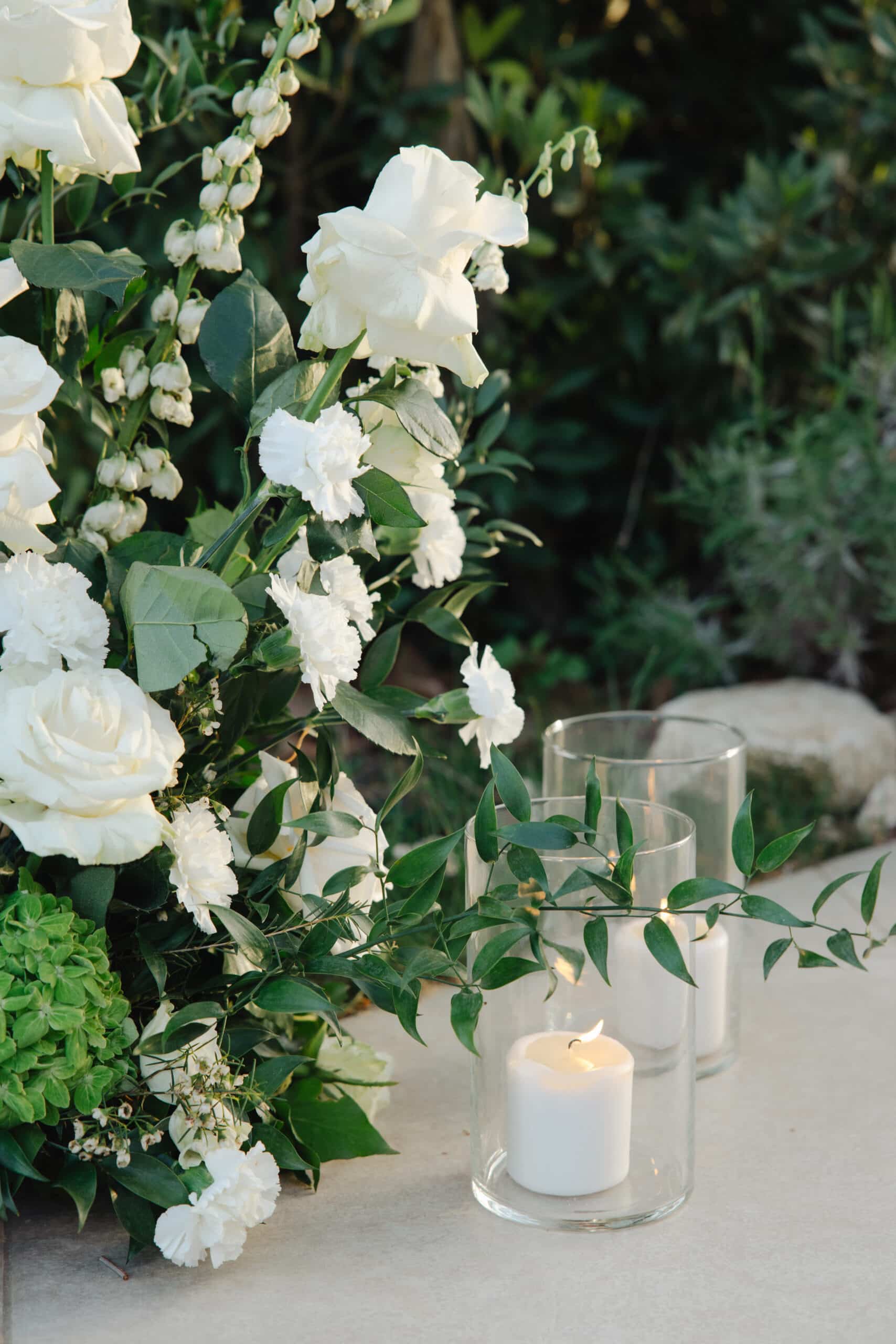 Gros plan d'un arrangement de roses et œillets blancs avec feuillage vert luxuriant. Deux bougies en verre éclairent la décoration.