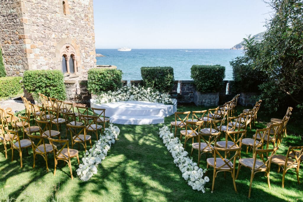 Cérémonie de mariage extérieure de luxe sur pelouse. Allée de fleurs blanches, chaises bois, scène face à la mer et mur de château historique.