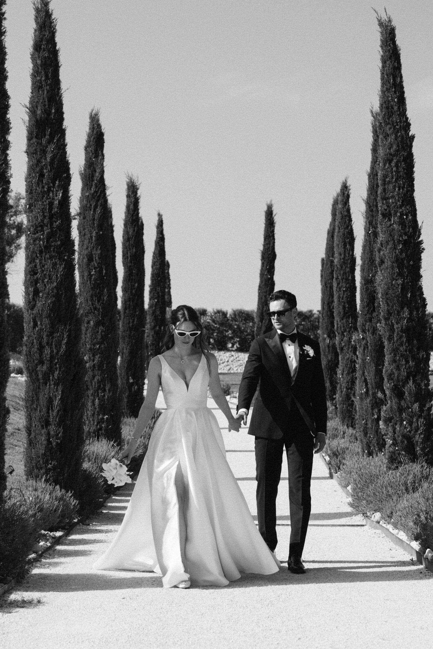 Mariés élégants en robe à fente et smoking, marchant main dans la main dans une allée de gravier bordée de cyprès sous le soleil.
