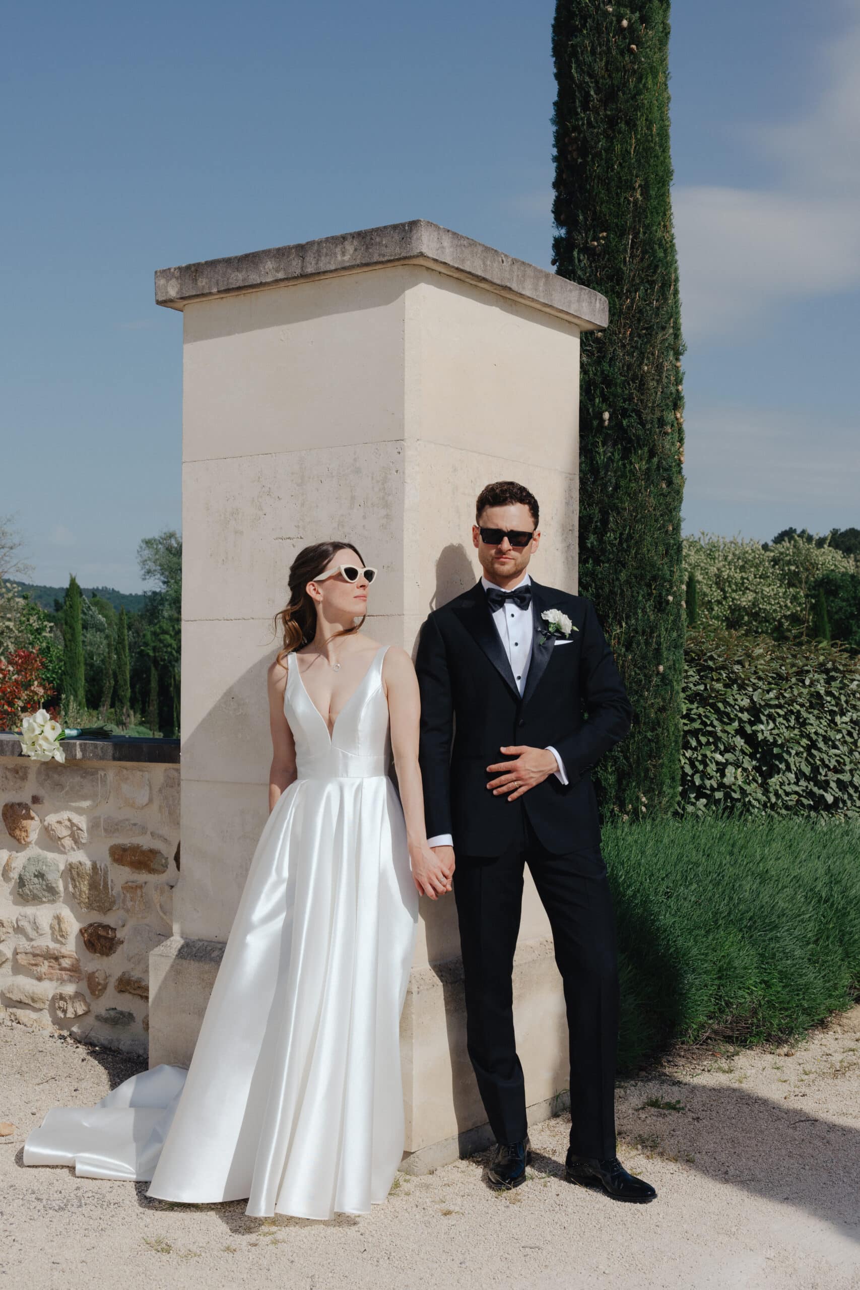 Jeunes mariés élégants en lunettes de soleil posant contre un pilier de pierre. La mariée porte une robe en satin, le marié un smoking noir.
