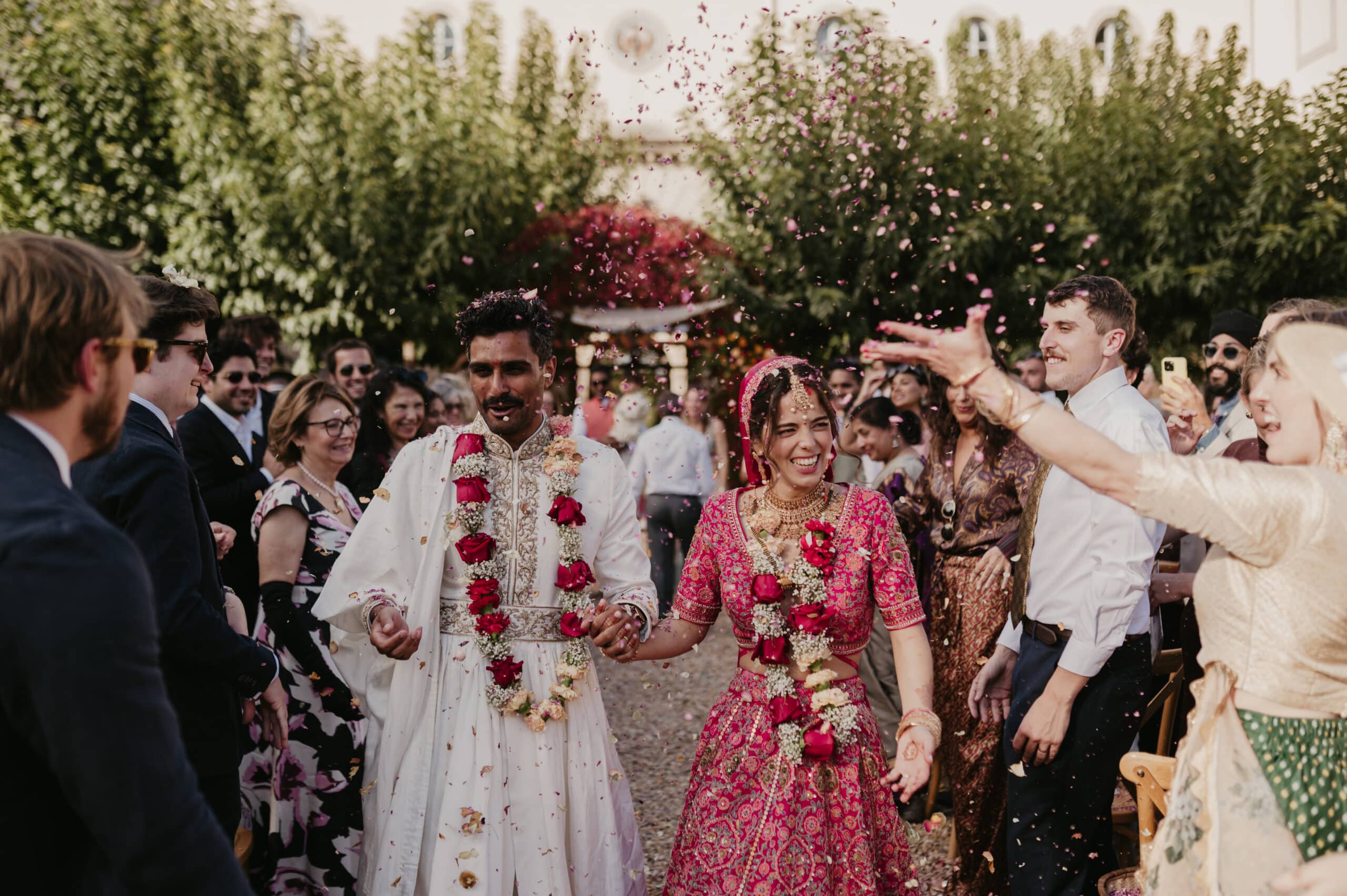 Mariage indien festif. Le couple, vêtu de tenues traditionnelles roses et blanches, sourit en marchant sous une pluie de pétales de fleurs.