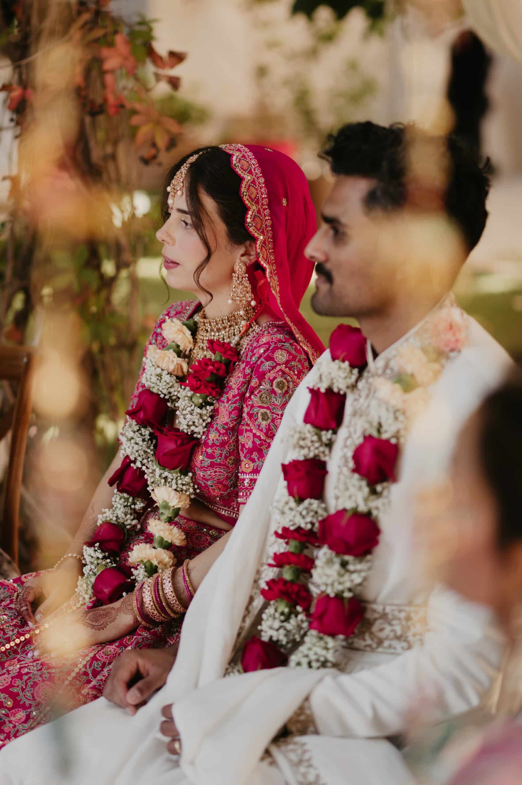 Cérémonie de mariage indien : mariée en lehenga rose vif brodé et bijoux traditionnels, assise à côté du marié en blanc. Varmalas de roses.