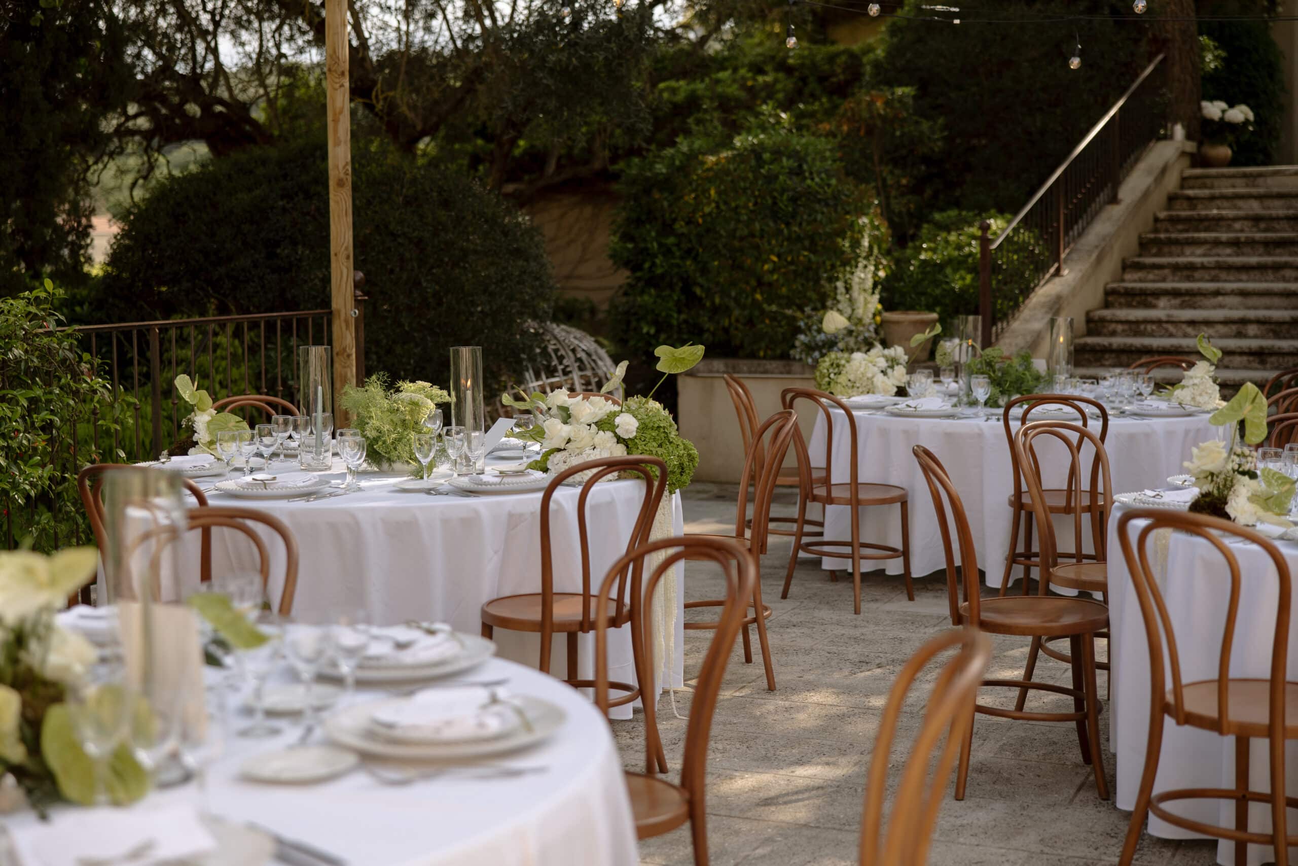 Réception de mariage élégante sur terrasse : tables rondes, chaises Thonet en bois, décorations florales blanches et verdure luxuriante.