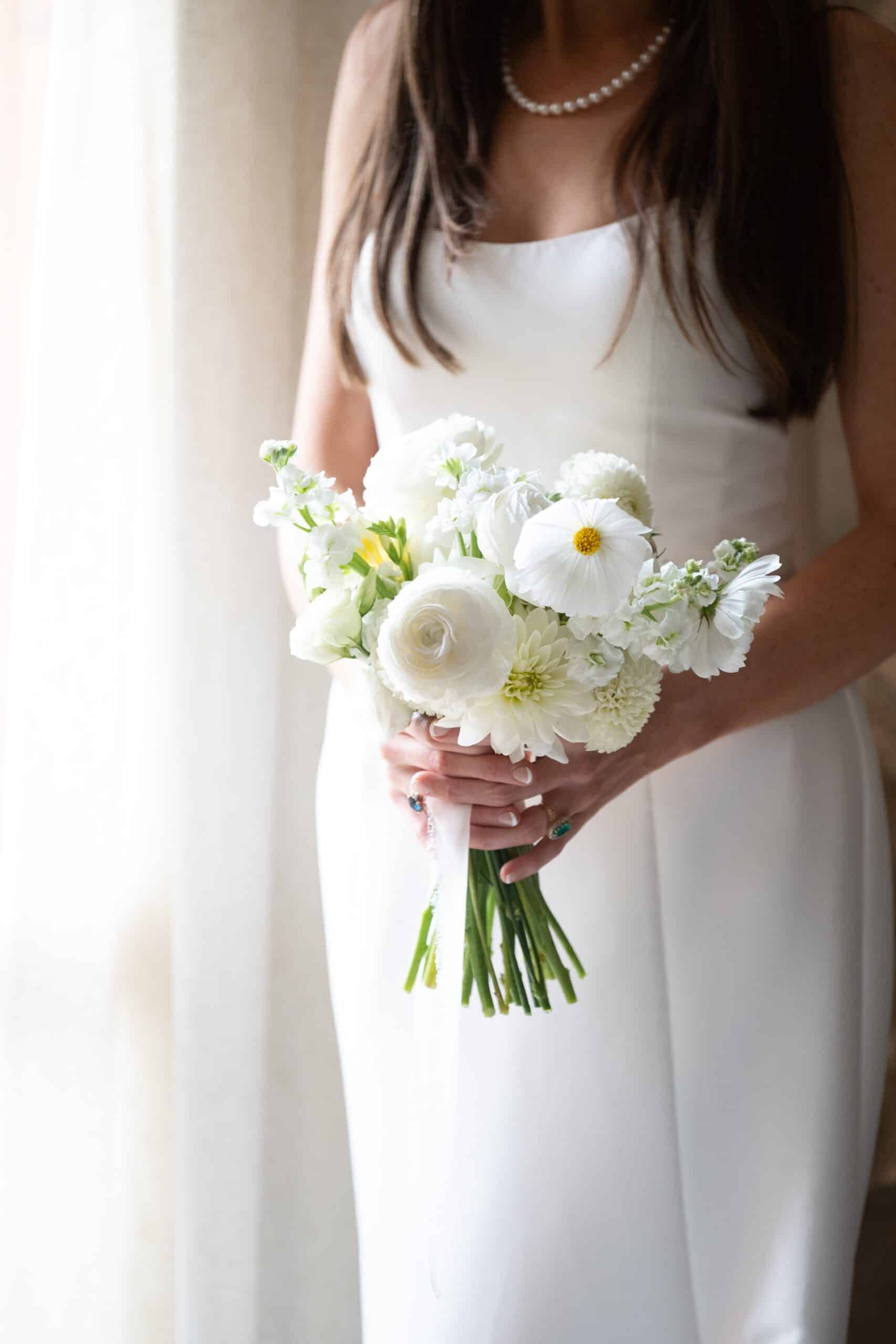 Mariée en robe blanche simple tenant fermement un bouquet de ranunculus et dahlias blancs, un collier de perles et des bagues aux mains.