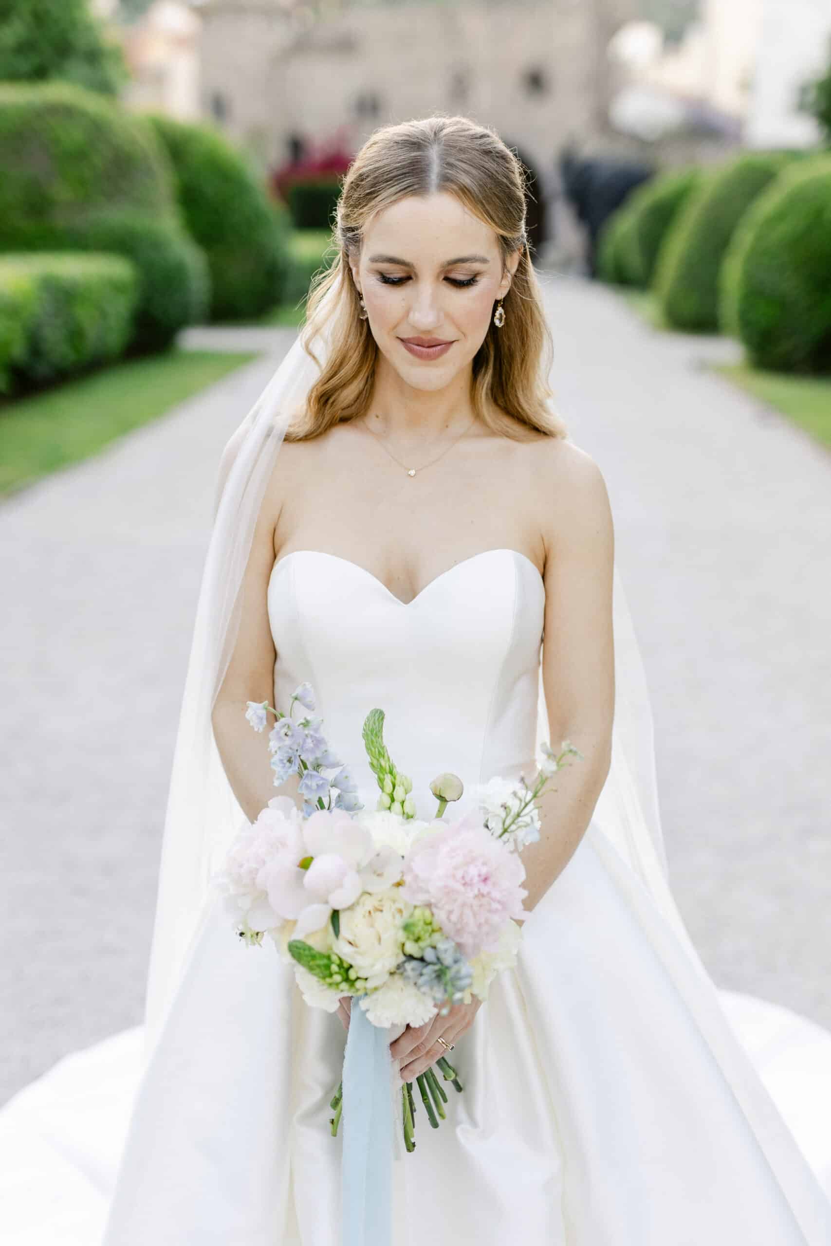 Belle mariée en robe bustier de satin blanc. Elle contemple son bouquet pastel sur l'allée d'un jardin classique.