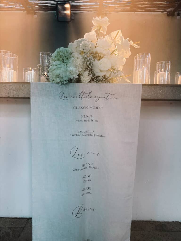 Menu Cocktails & Vins sur tissu lin : Mojito, Punch, Jacquelin, Tariquet, Grezan. Décor floral blanc romantique, lumières douces.
