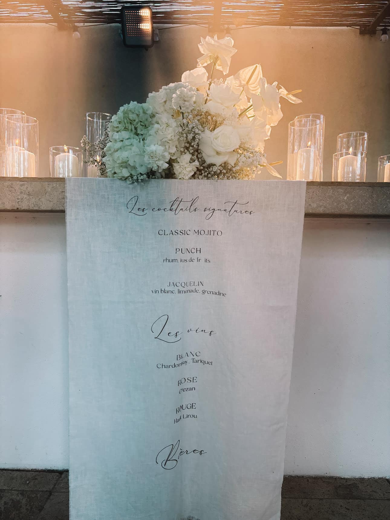 Menu Cocktails & Vins sur tissu lin : Mojito, Punch, Jacquelin, Tariquet, Grezan. Décor floral blanc romantique, lumières douces.
