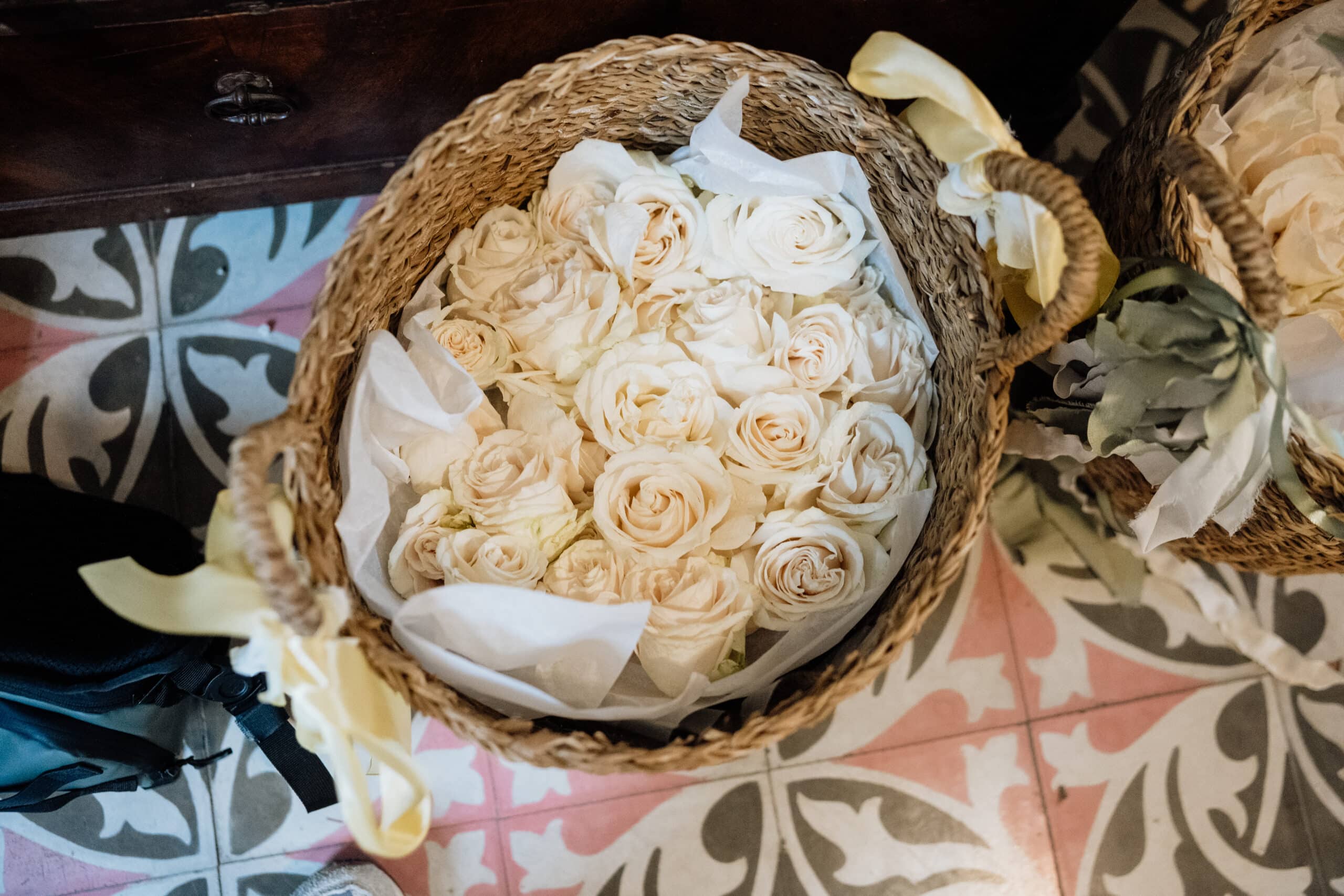 Panier d'osier rempli de roses ivoires et papier blanc, posé sur des carreaux de ciment anciens à motifs floraux roses et gris.