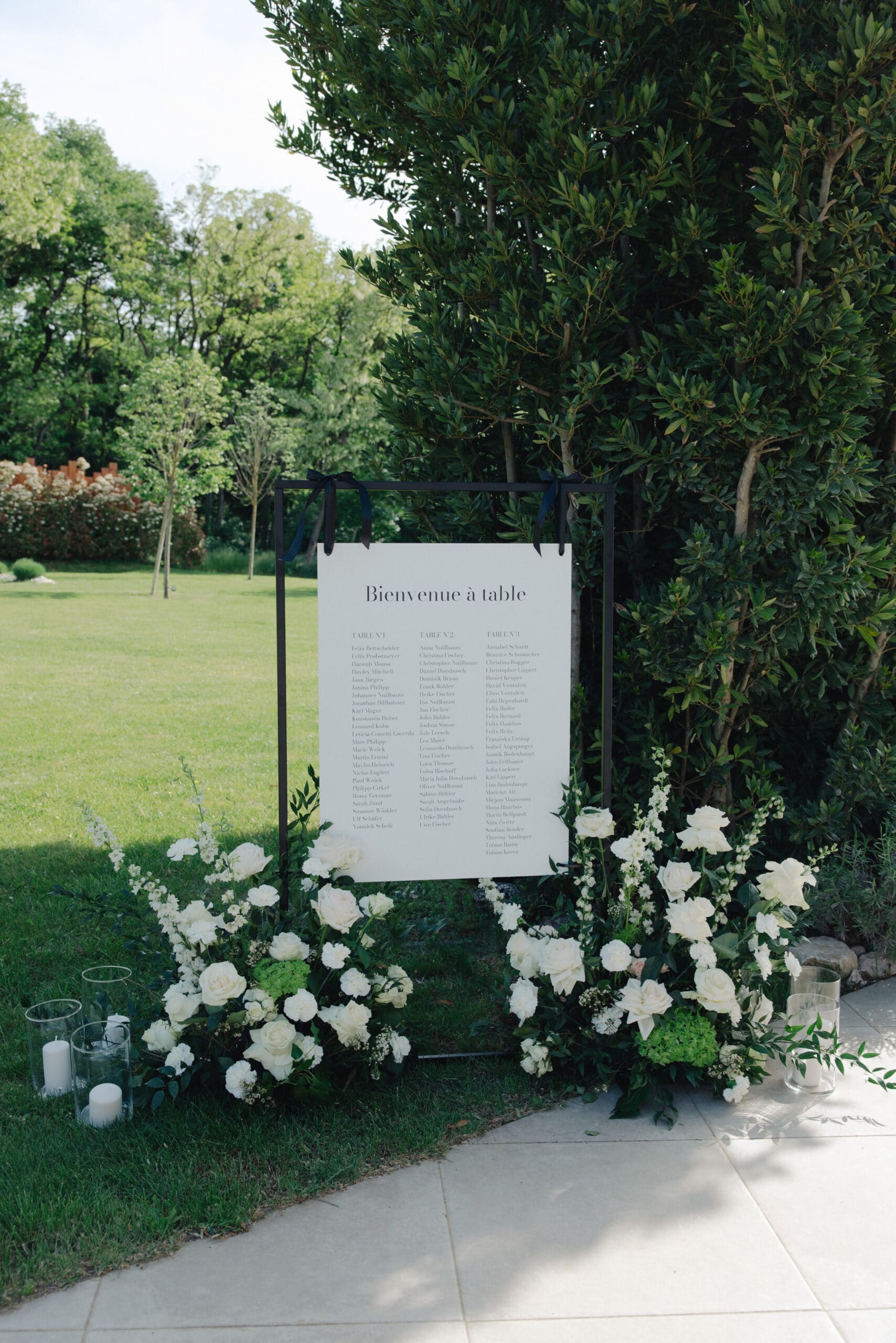 Panneau de placement "Bienvenue à table" en français, encadré par des haies. Décor de mariage élégant : roses blanches et bougies sur pelouse.