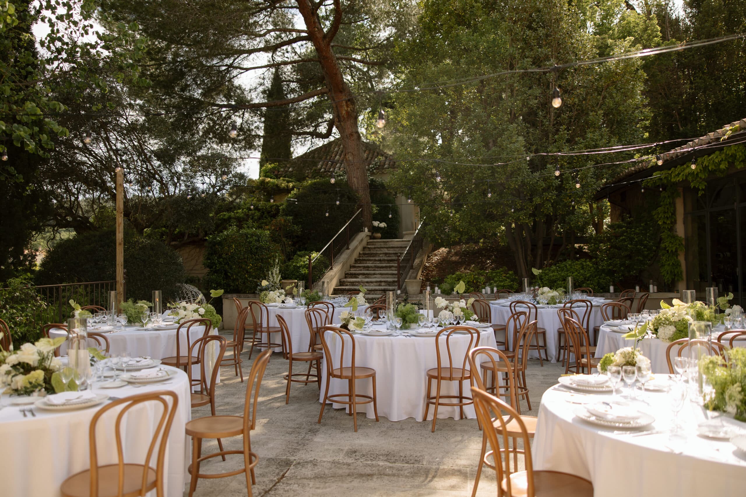 Tables rondes dressées pour une réception de mariage en plein air. Chaises Thonet, décorations florales blanches et escalier en pierre du jardin.
