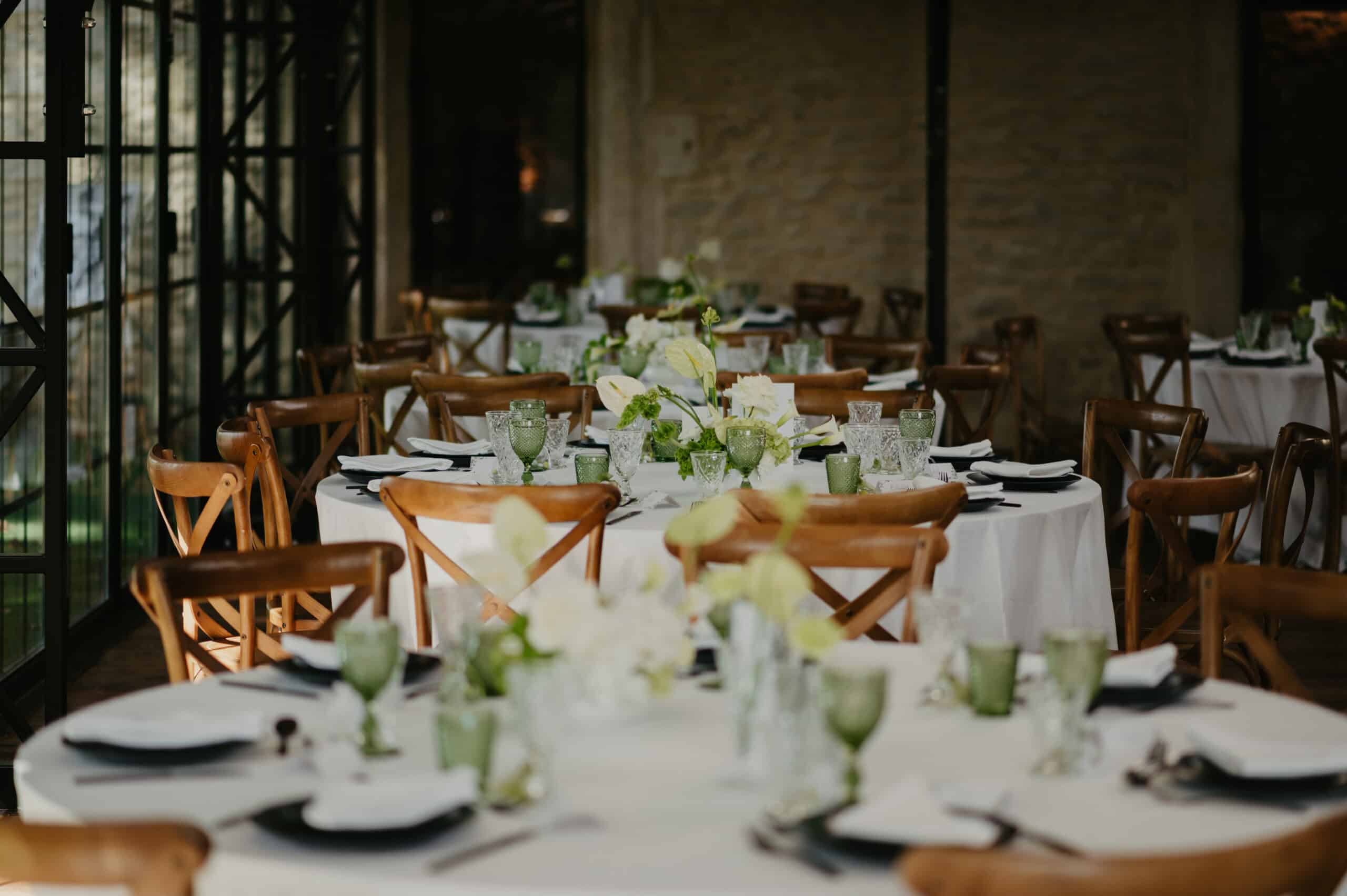 Salles de réception rustique. Tables dressées de nappes blanches, chaises en bois X, et centres floraux blancs et verts. Verres verts texturés.