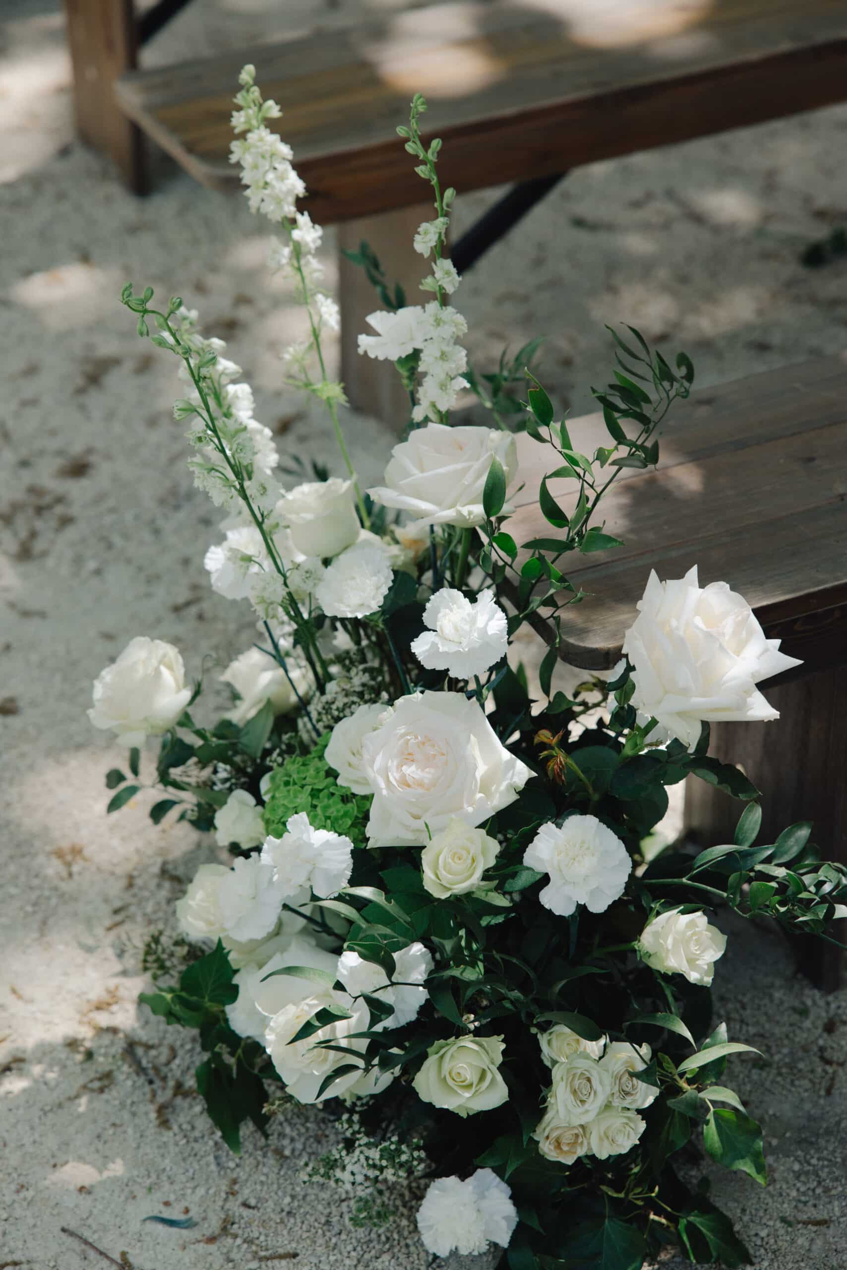 Arrangement floral extérieur luxueux : roses et œillets blancs éclatants, haute verdure et delphiniums, devant des bancs rustiques.