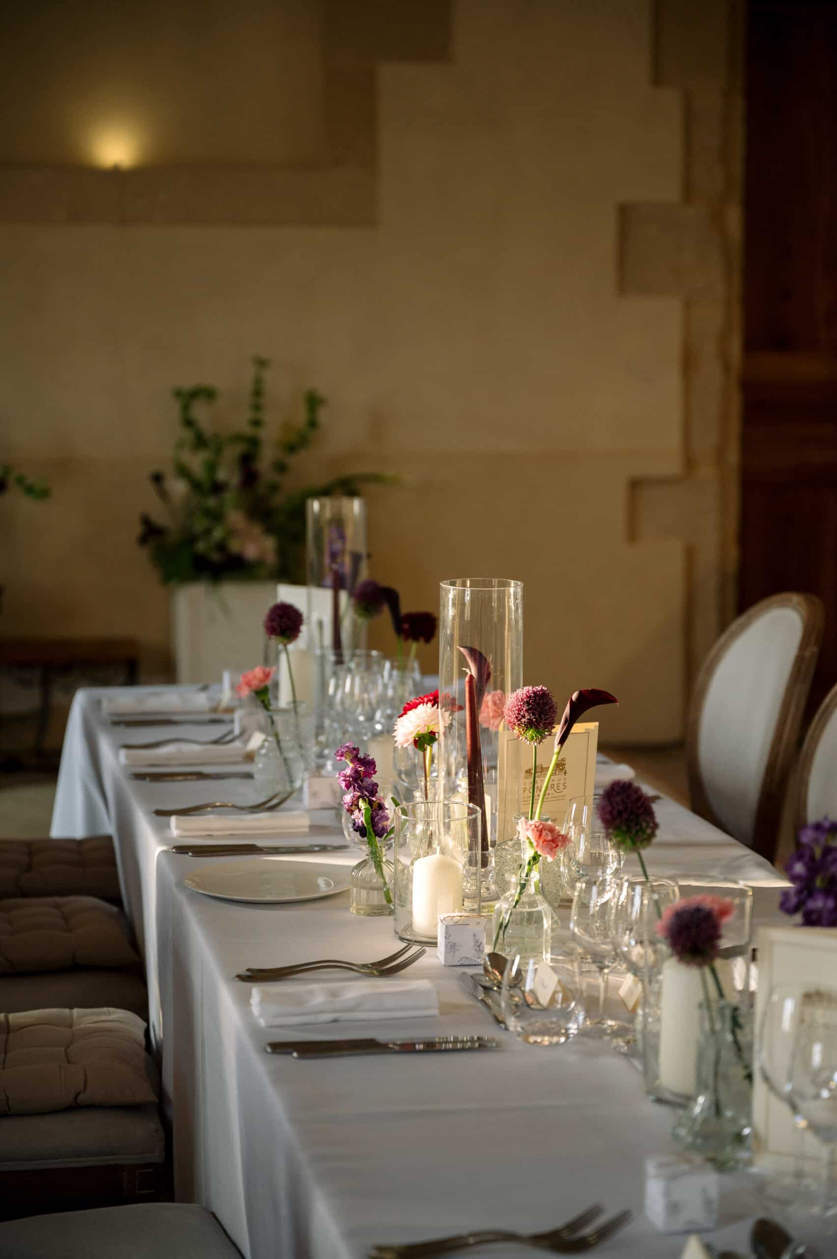 Table de dîner élégante dressée avec nappe blanche et couverts. Décoration florale de mariage (alliums, callas) et bougies. Ambiance chaleureuse.