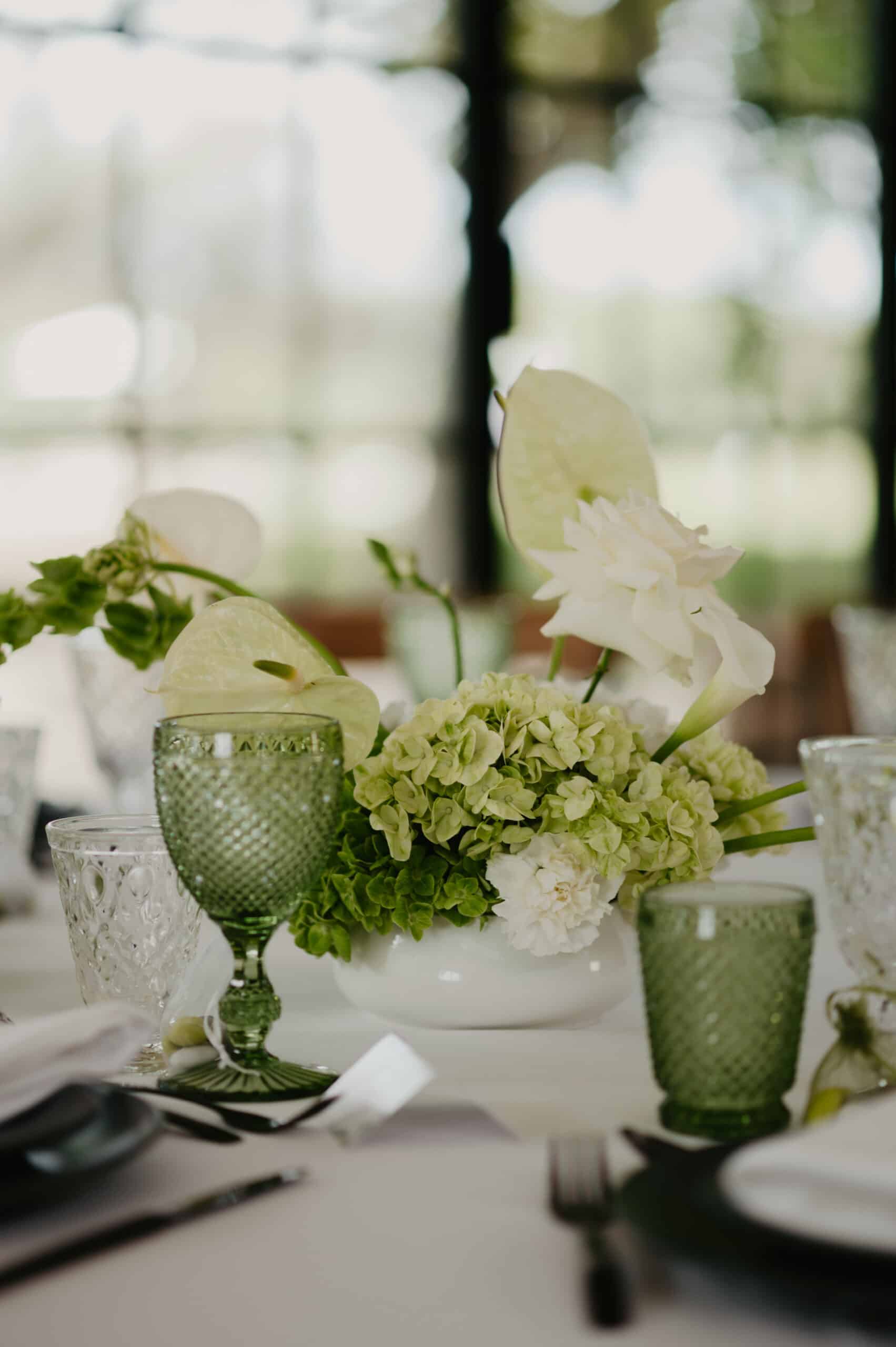 Centre de table vert et blanc pour mariage, composé d'hydrangées, d'anthuriums et de roses. Verrerie verte texturée sur nappe blanche.